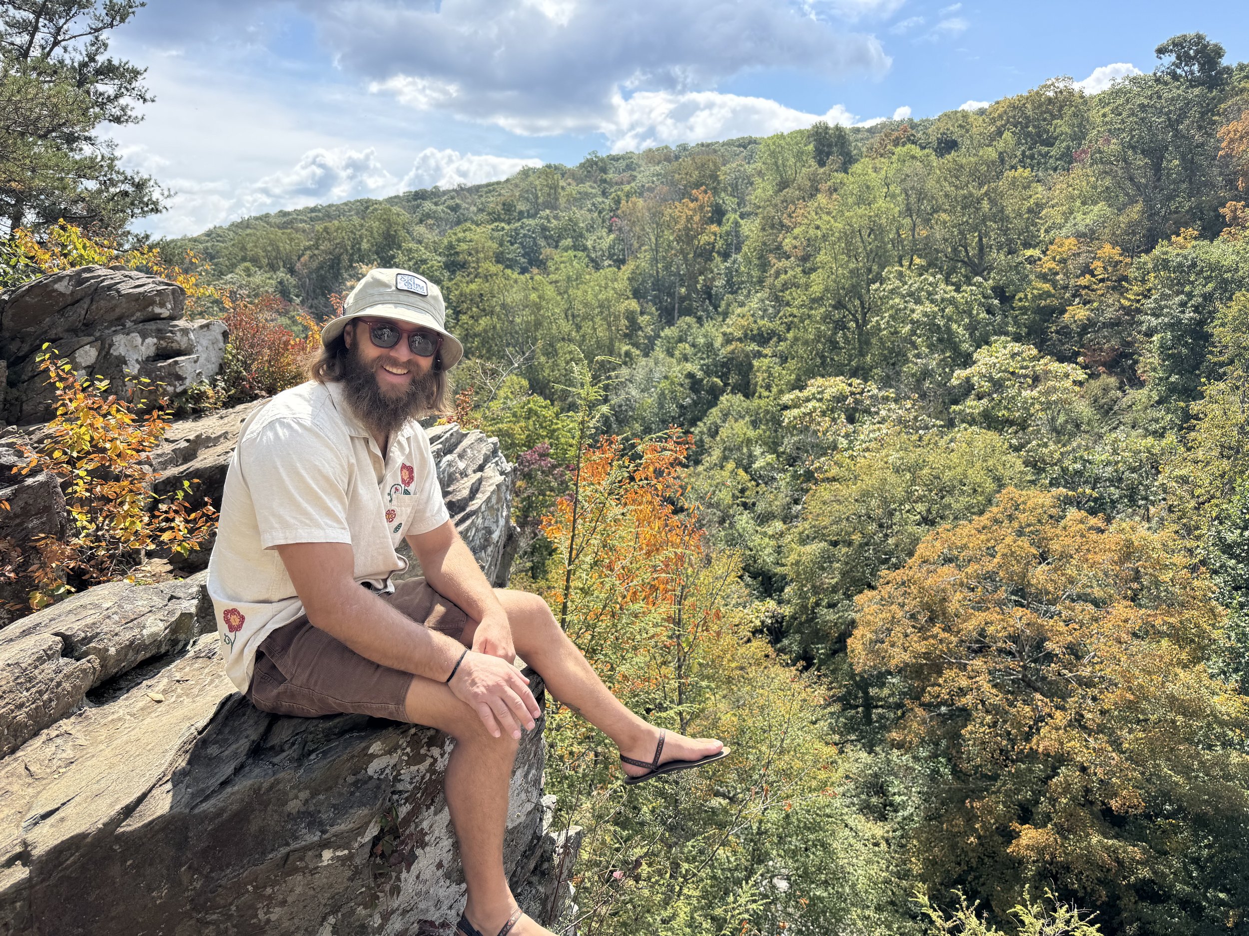 A man with a beard, wearing sunglasses, a beige hat, and a light-colored shirt, is sitting on a rocky ledge in a forested area during fall, with green and orange leaves, under a partly cloudy sky.