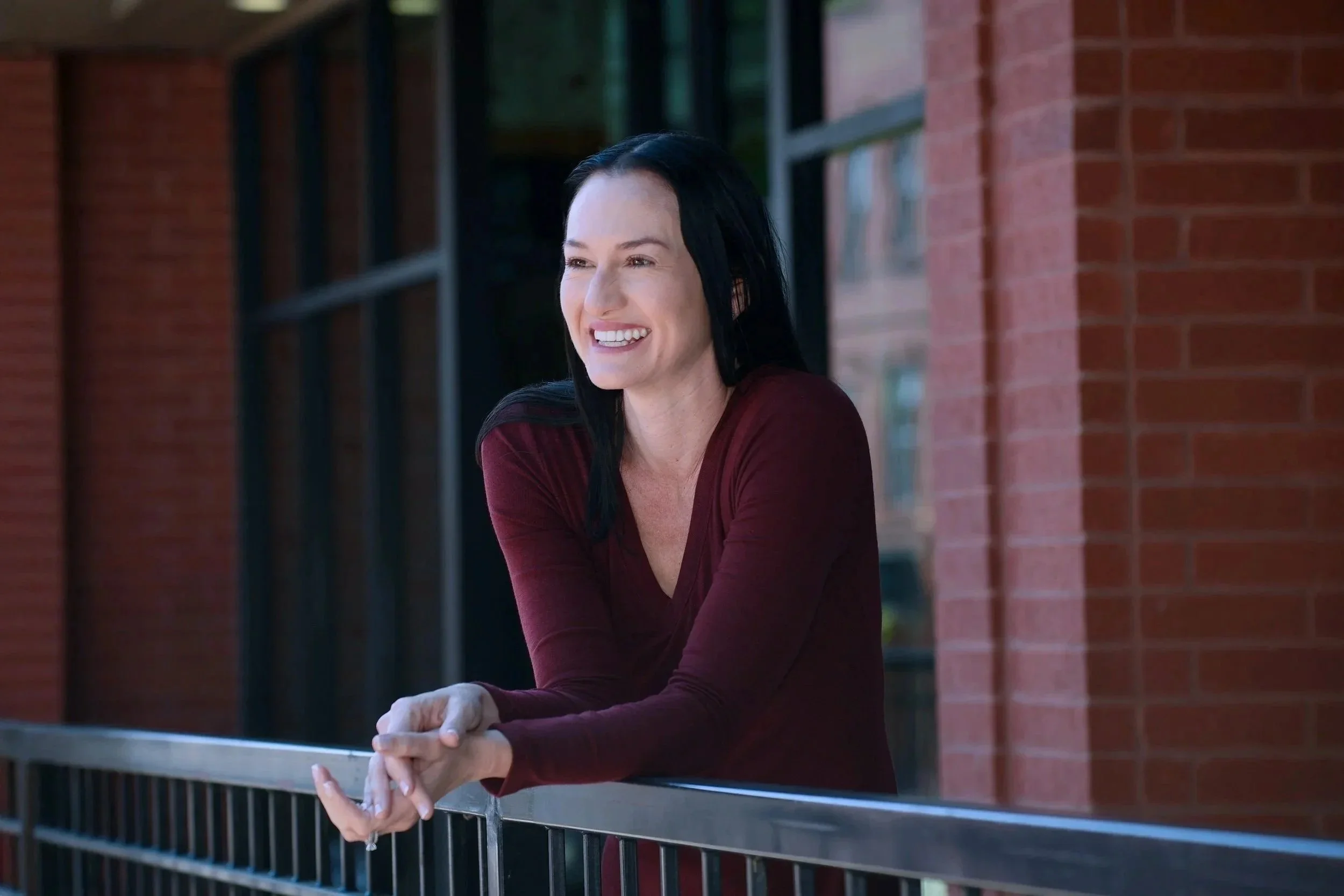 A woman with long black hair smiling and leaning on a metal railing outside a brick building. She is wearing a burgundy long-sleeve top.