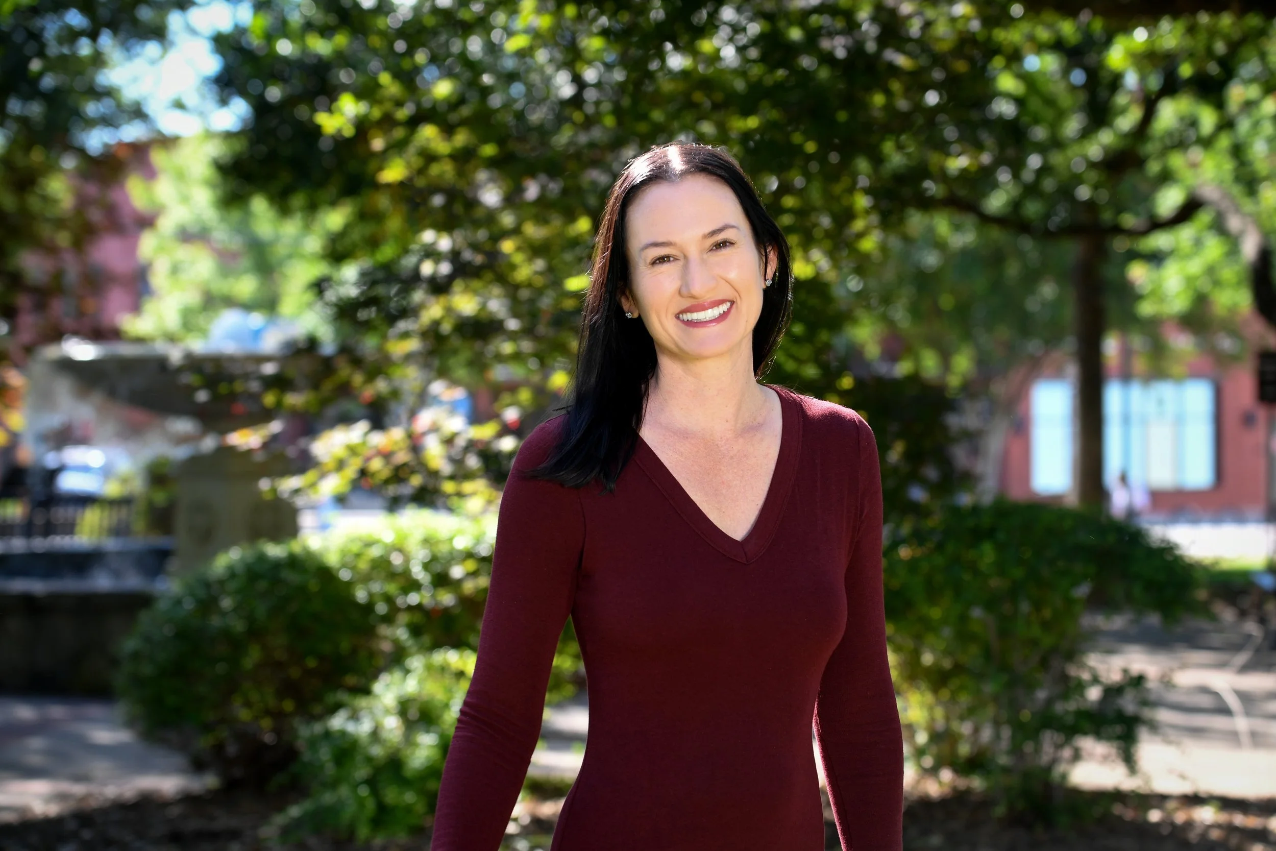 A woman with long black hair smiling outdoors in front of green trees, wearing a maroon long-sleeve top.