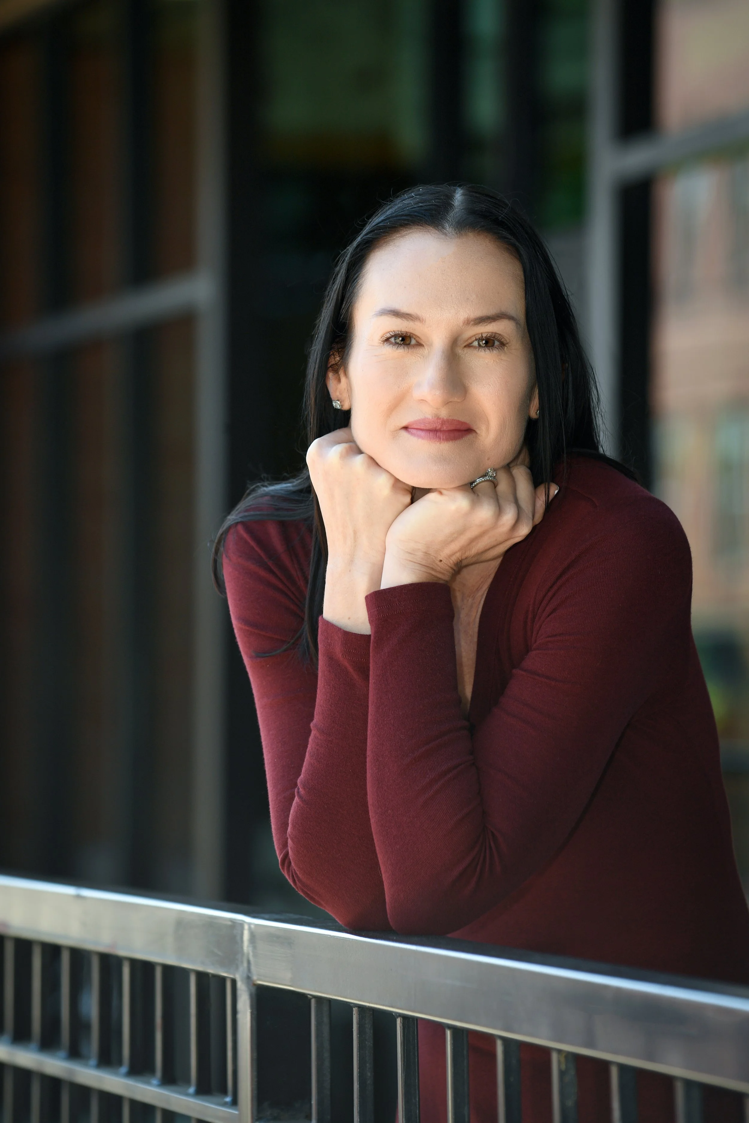 A woman with dark hair and light skin, resting her chin on her hands, leaning on a metal railing, wearing a maroon long-sleeve shirt, outdoors near a building with large windows.