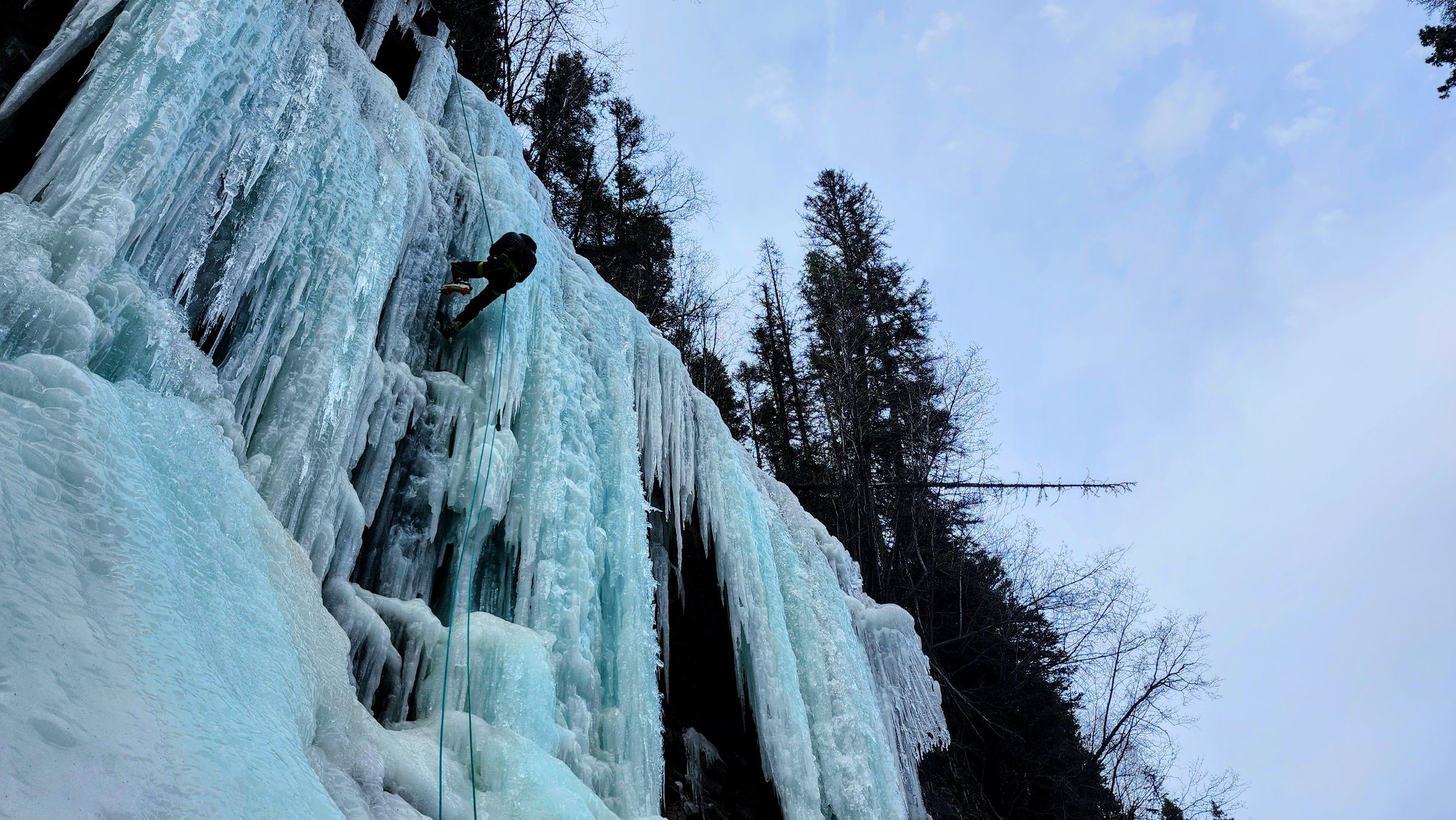 A person wearing winter gear climbing a frozen icy waterfall with trees in the background.