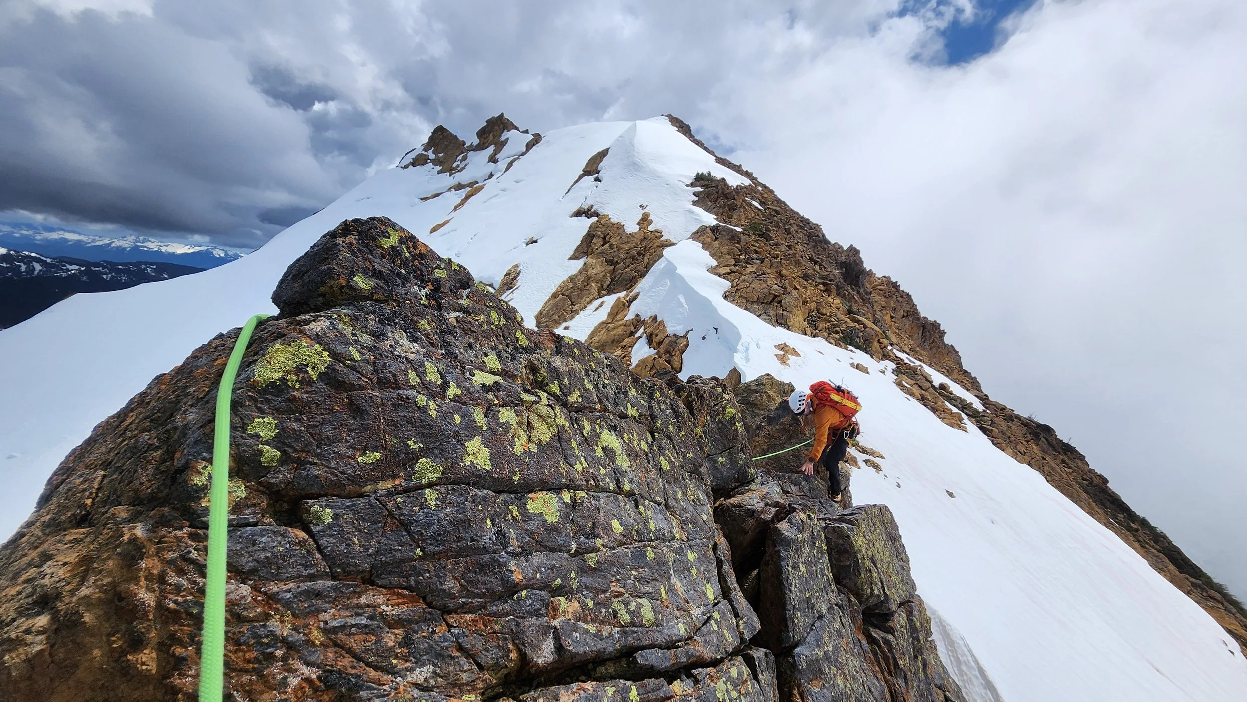 A person wearing an orange jacket, helmet, and climbing gear ascending a rocky, snowy mountain ridge with clouds and distant snow-capped peaks in the background.
