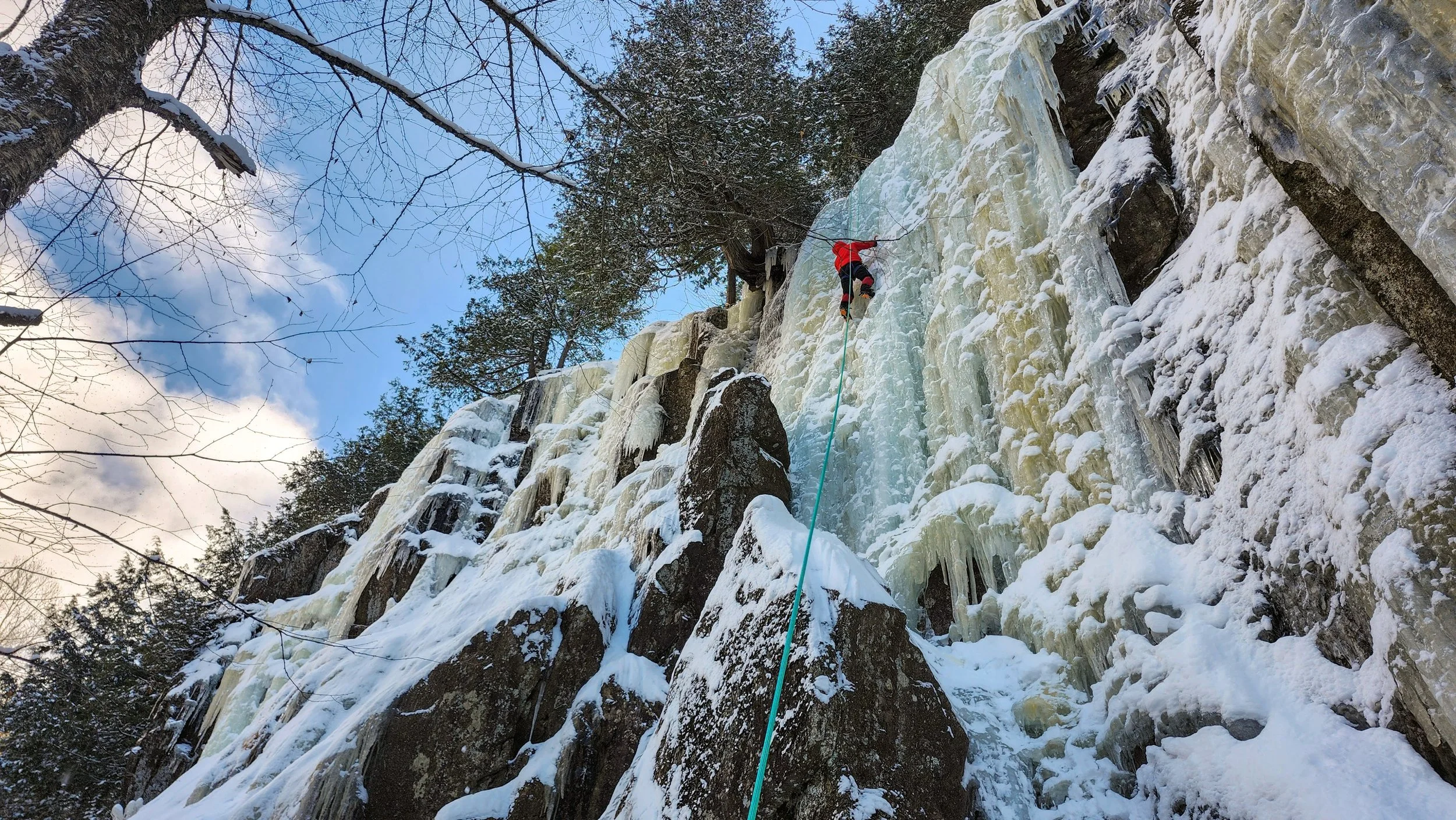 A person ice climbing on a frozen waterfall, wearing a red jacket and black pants, with a blue rope securing them, surrounded by snow and ice, with a sky and leafless trees overhead.