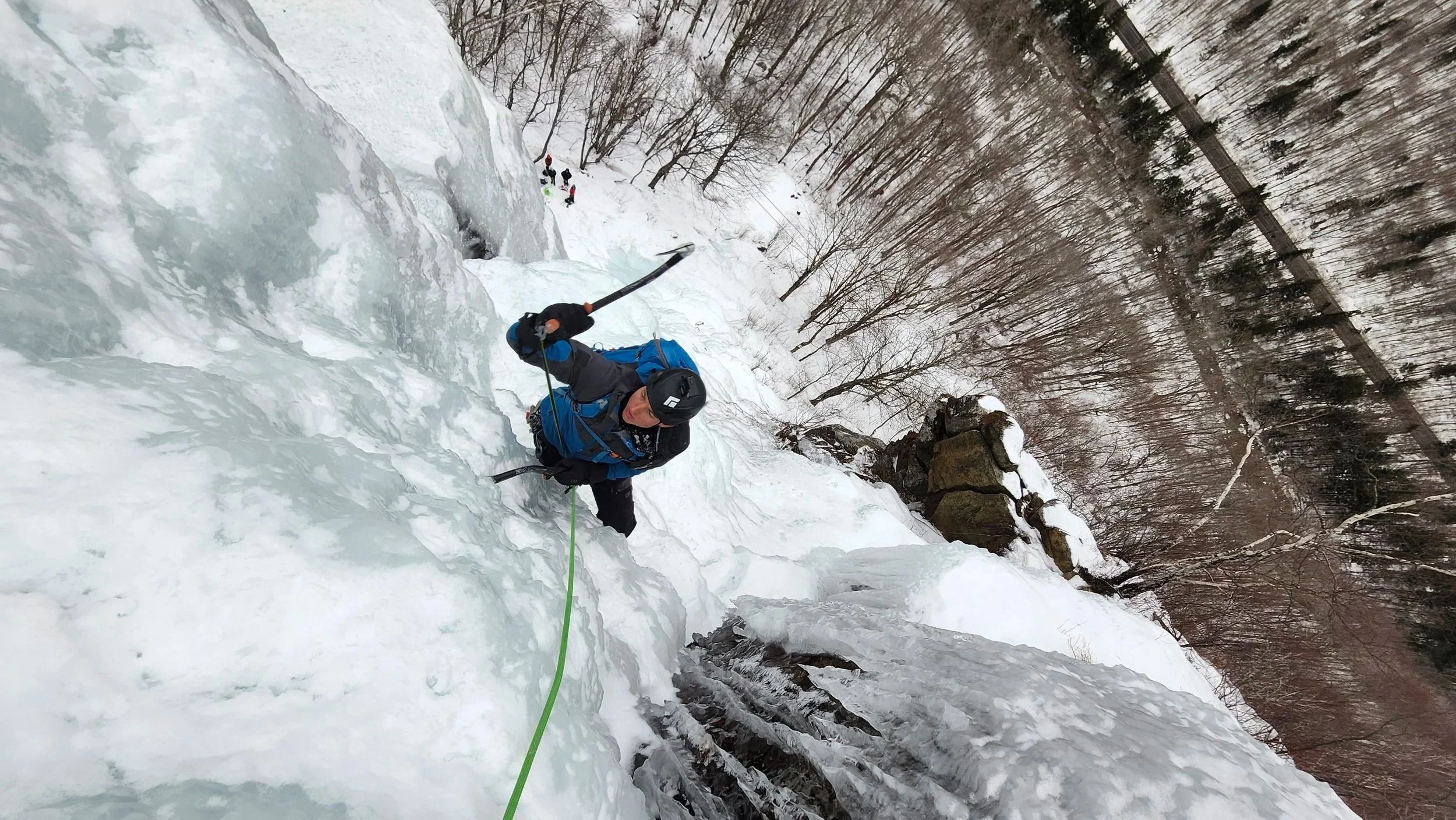 A person wearing a helmet and outdoor gear is climbing a frozen waterfall, using an ice axe and ropes, with a snowy forest in the background.