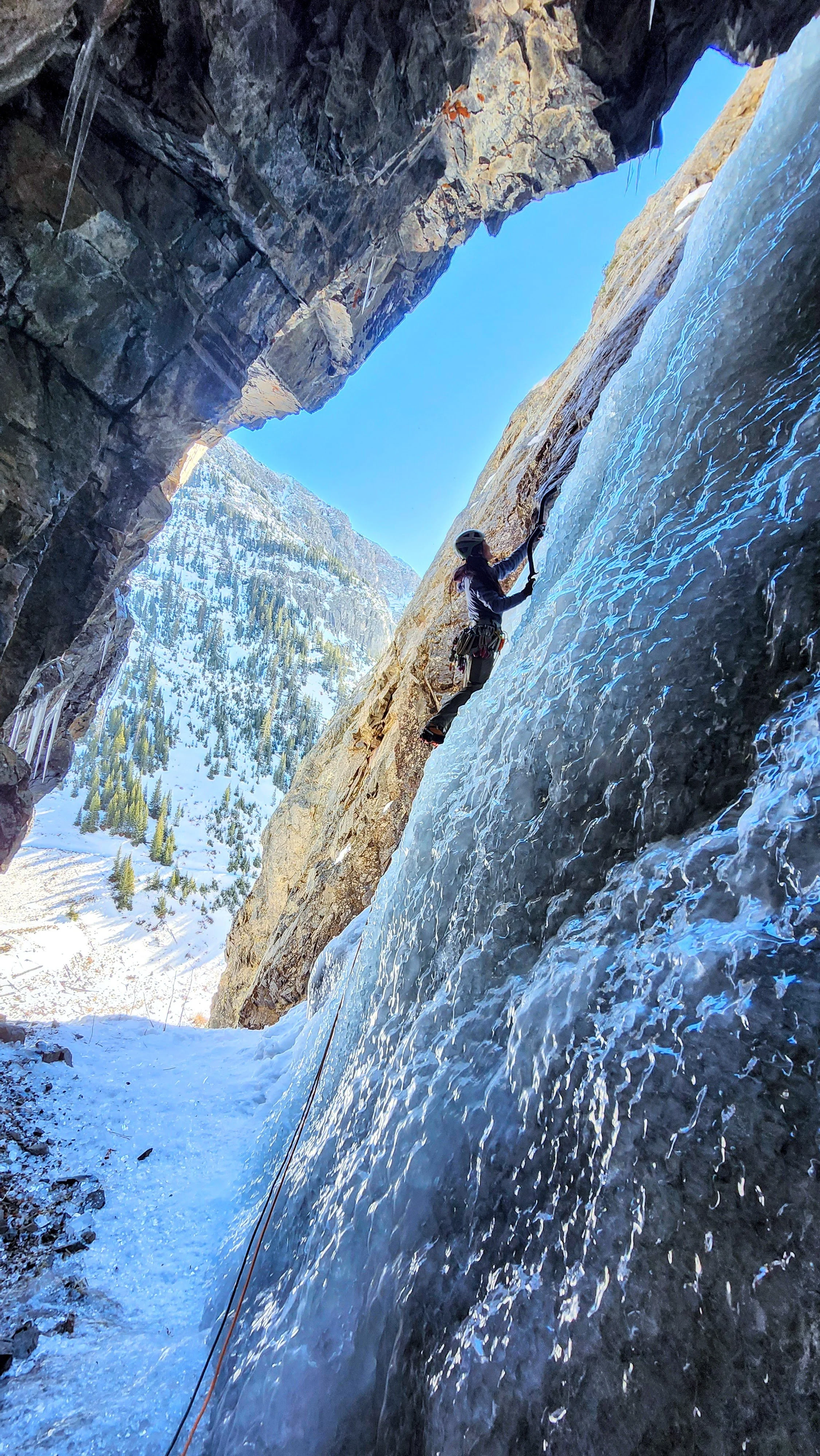 A person ice climbing on a frozen waterfall in a snowy mountainous landscape, wearing winter gear and a helmet, with snow-covered trees in the background.