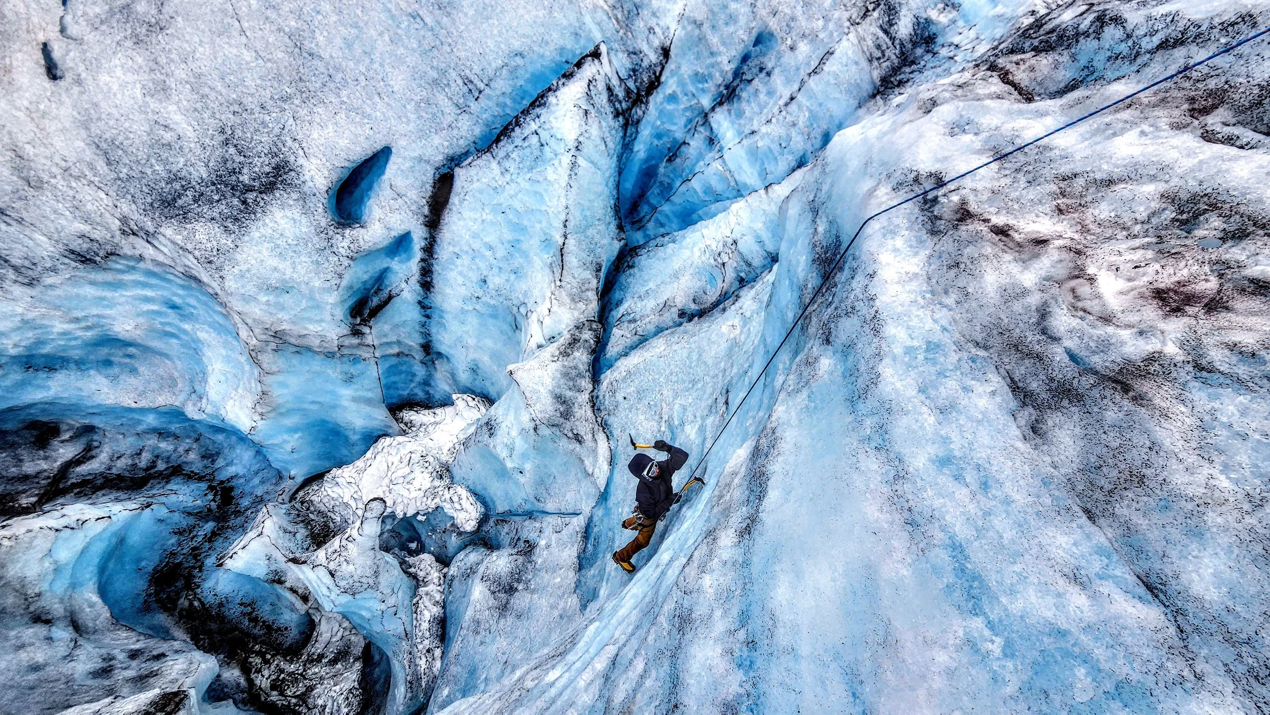 A person in outdoor gear ice climbing on a glacier with blue ice and snow, secured by a safety rope.