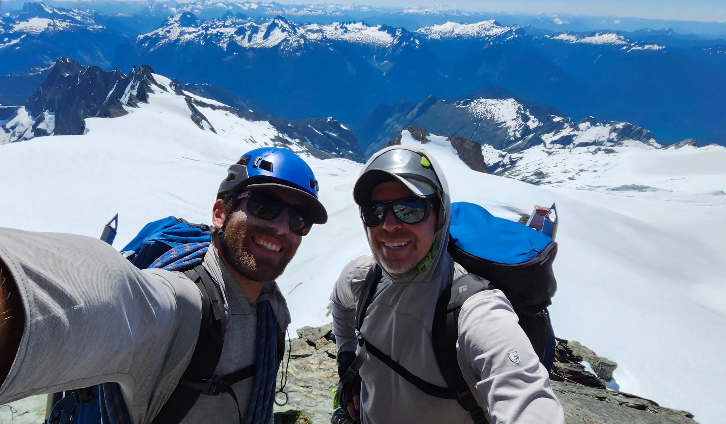 Two smiling men wearing helmets and sunglasses taking a selfie on a snowy mountain with a mountain range in the background.