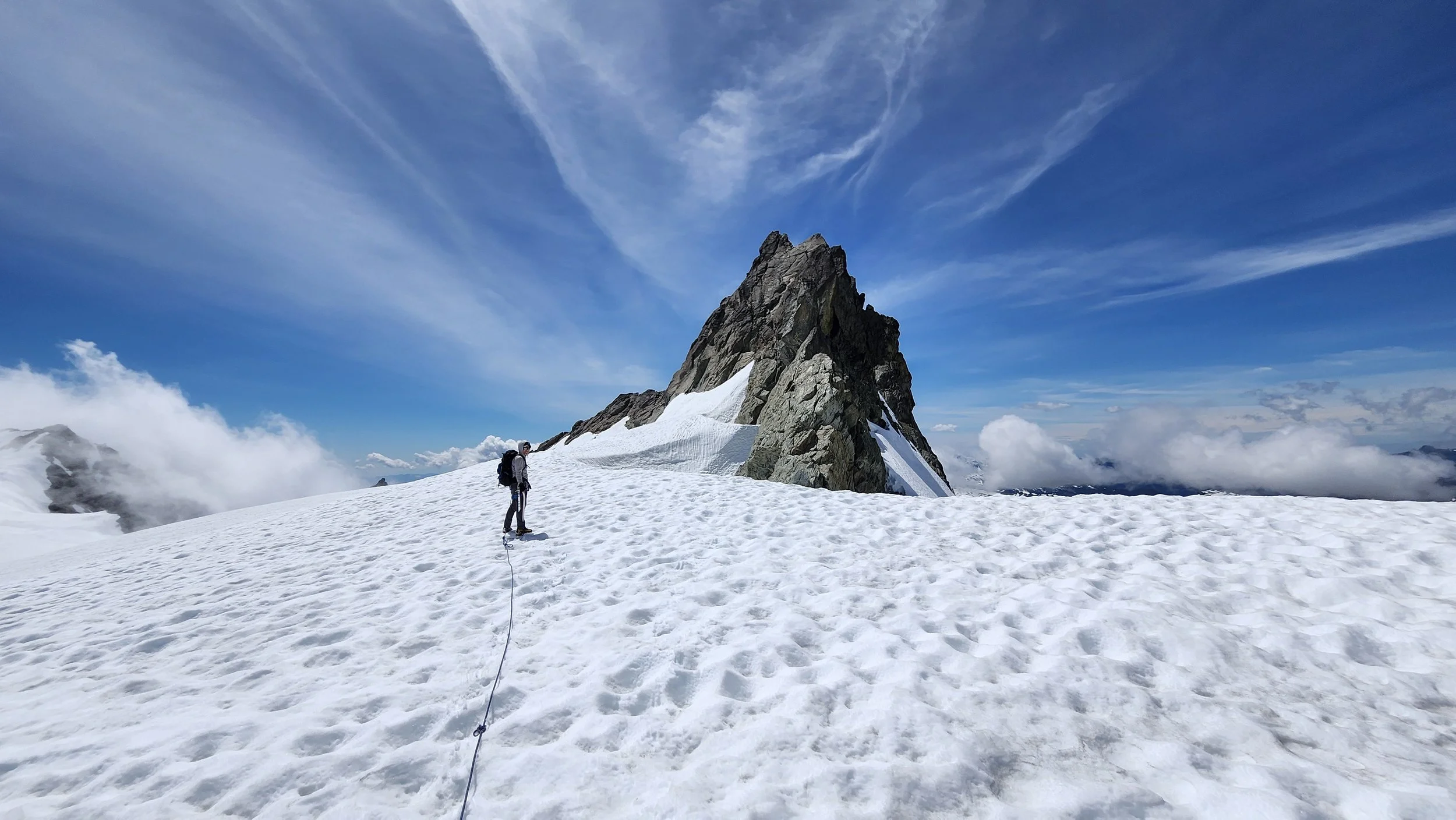 A person climbing a snowy mountain with a large rocky peak in the background under a blue sky with wispy clouds.