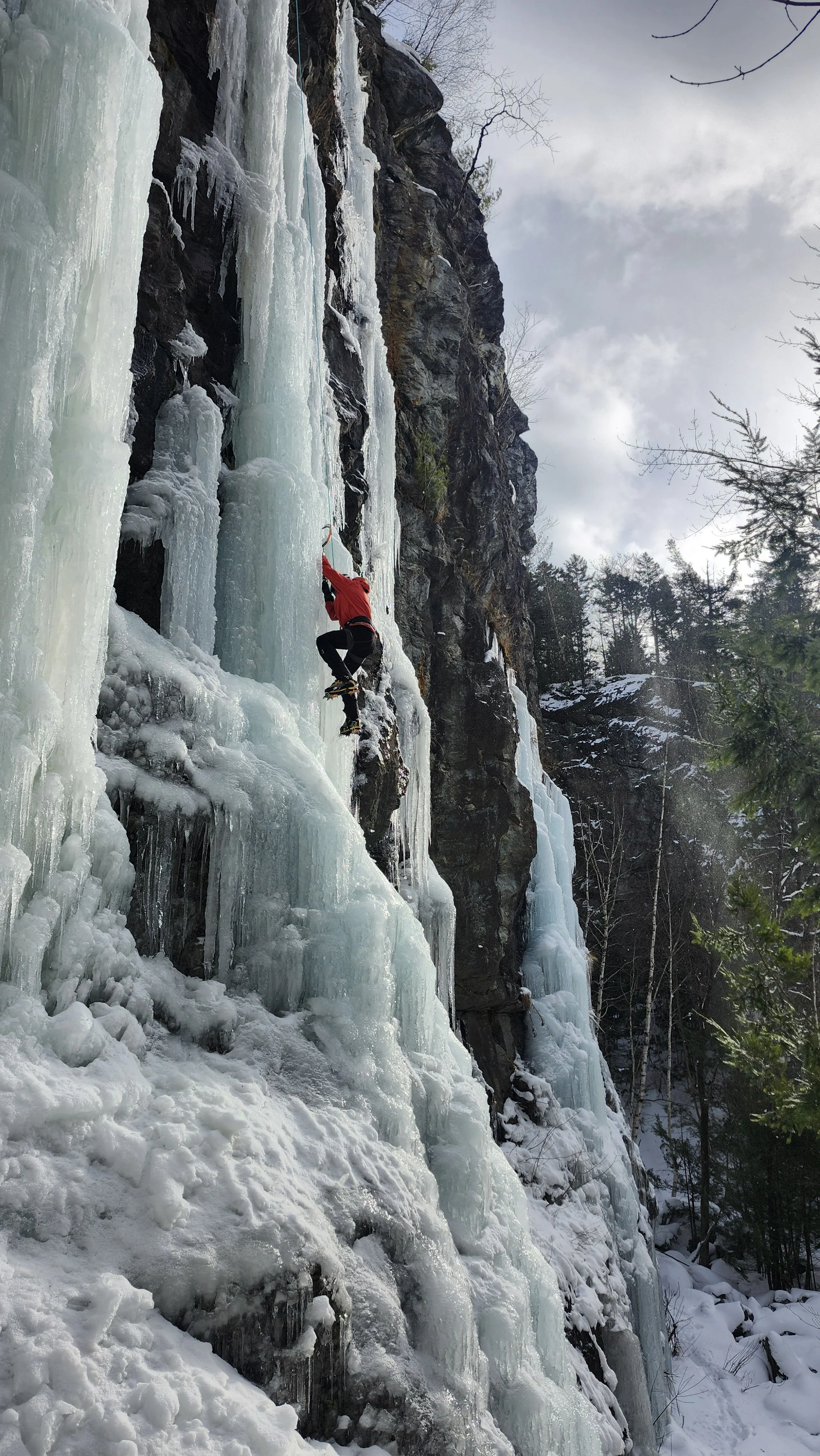 A person wearing a red jacket and black pants ice climbing on a frozen waterfall with icicles and snow-covered rocks and trees in the background.