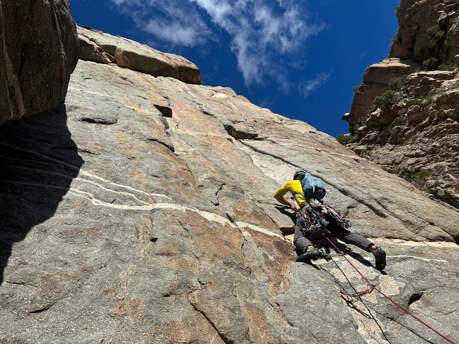 A person rock climbing on a steep cliff face under a blue sky, wearing a yellow shirt, helmet, and climbing gear.