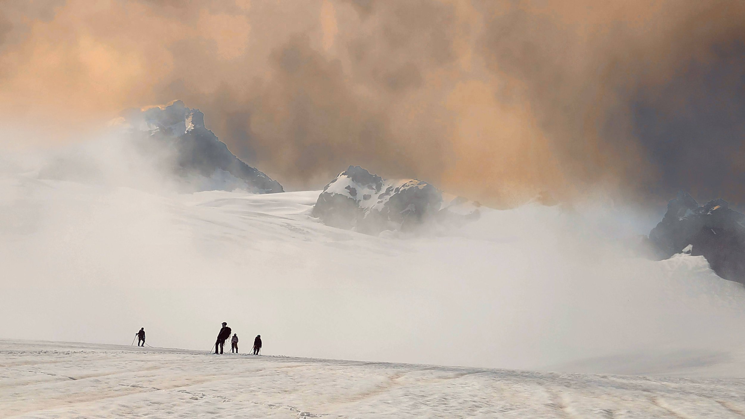 A group of five hikers walking across a snow-covered landscape toward distant mountain peaks shrouded in clouds and mist.
