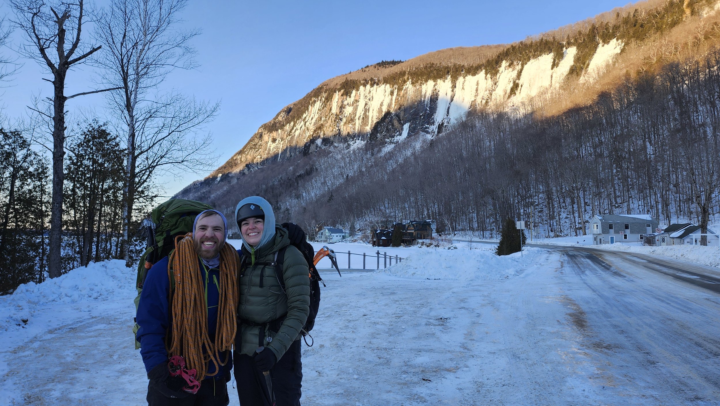 Two smiling hikers with backpacks standing outdoors in a snowy rural area, with a mountain and a few houses in the background during late afternoon or early evening.