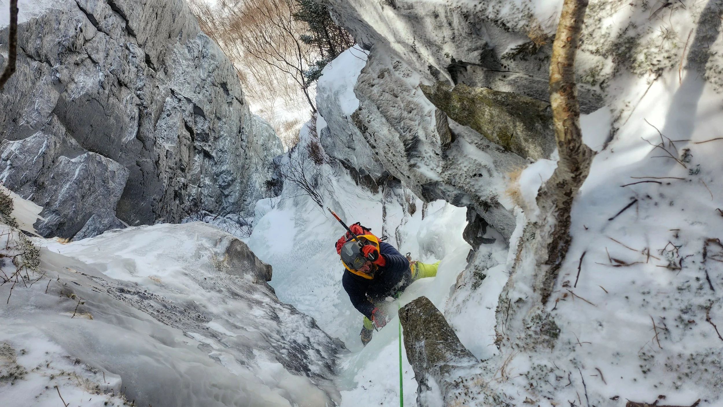 A person in cold-weather gear ice climbing up a narrow, snowy canyon with steep rock walls on either side.