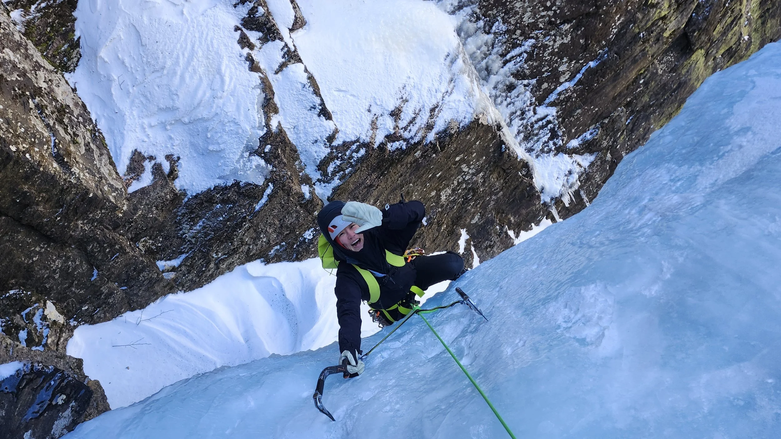 A person ice climbing on a snowy, icy cliff face with ice tools, wearing a helmet and winter gear, smiling and making a gesture with their hand, set against a mountainous landscape.