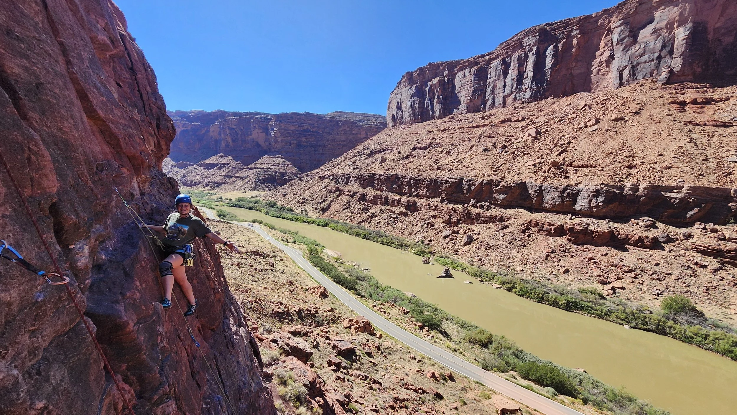 A woman in climbing gear and helmet comfortably resting on a steep mountain face with a scenic canyon landscape below, including a river, winding road, and rugged red rock cliffs under a clear blue sky.
