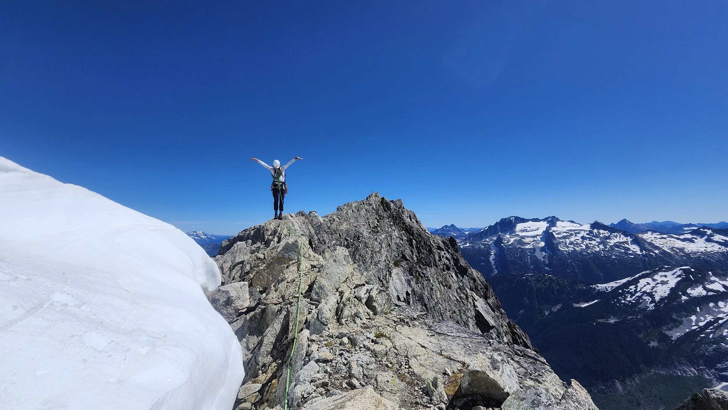 A person standing on a rocky mountain peak with arms outstretched, surrounded by snow and mountain ranges under a clear blue sky.