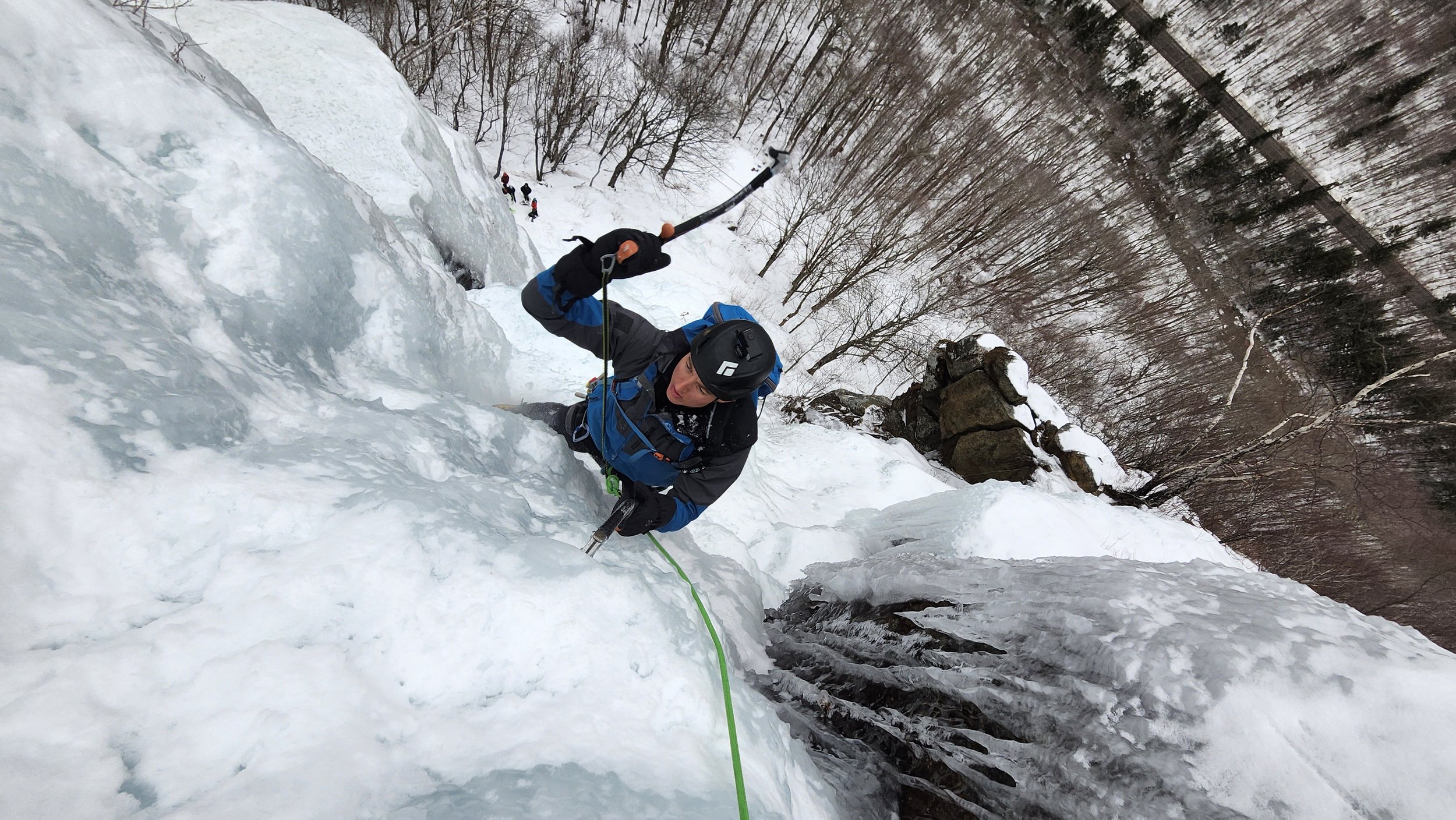 A person in mountaineering gear ascending an icy icefall with an ice axe, ropes, and winter clothing, surrounded by snow and leafless trees.