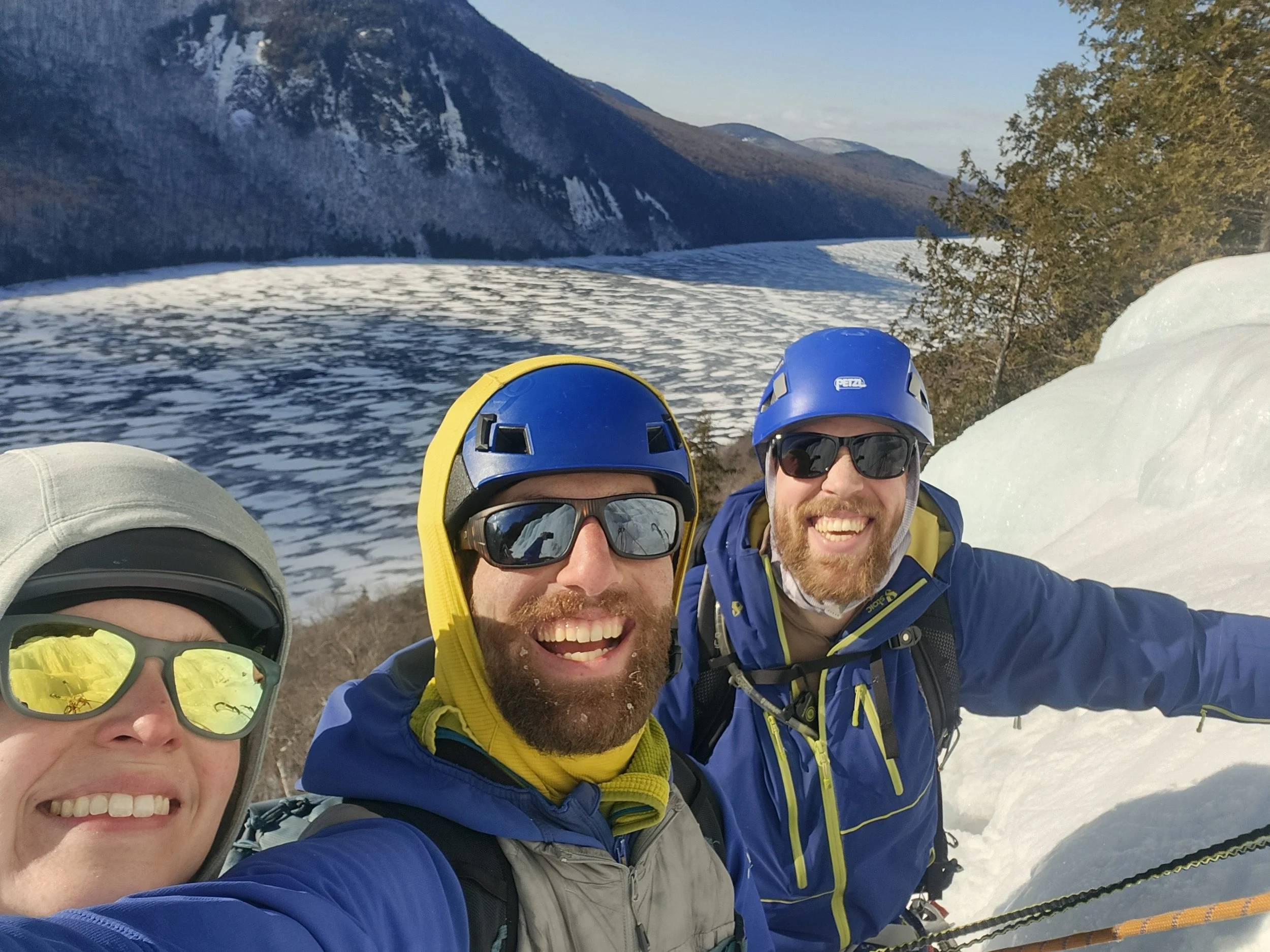 Three people wearing ski helmets and goggles smiling for a selfie in a snowy mountain landscape with trees and distant mountain slopes.