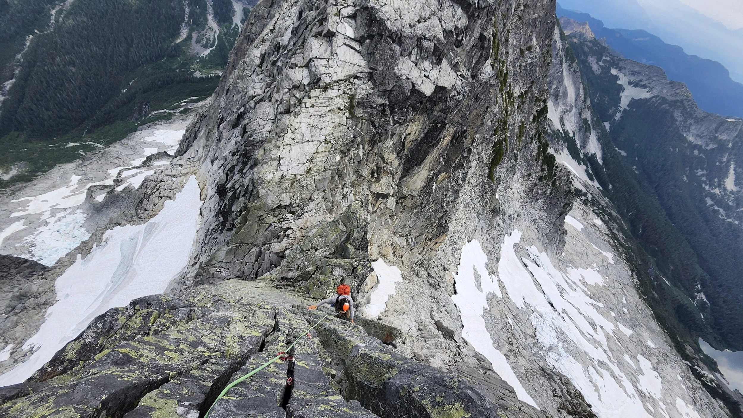 A person wearing an orange helmet and gray clothing is climbing a steep rocky mountain with snowy patches, using climbing gear and a safety rope, in a rugged mountain landscape.