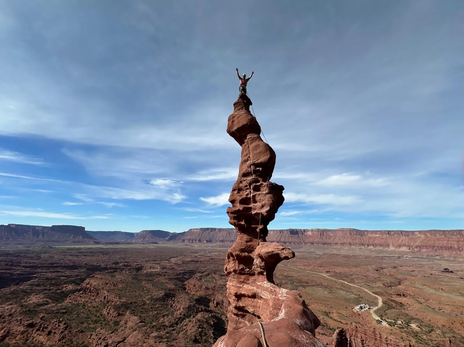 A person standing on top of a narrow, tall, and irregularly shaped rock formation in a desert landscape, with arms raised in victory or joy, and a vast canyon with layered red rock cliffs in the background.