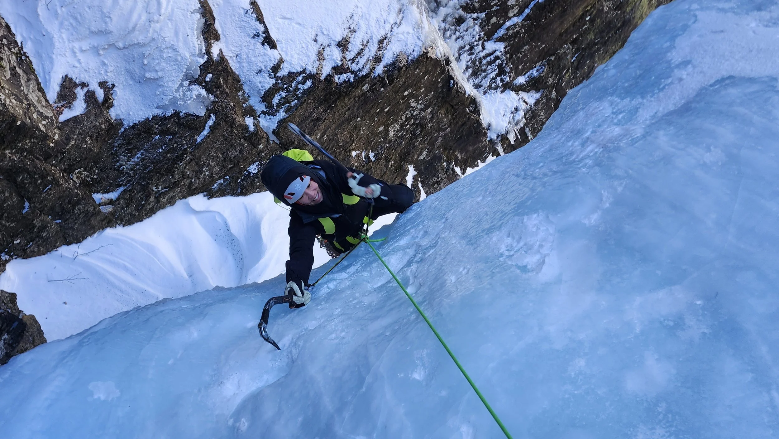 A person ice climbing on a frozen waterfall or ice formation, using climbing gear and a green rope, surrounded by rocky cliffs and snow.