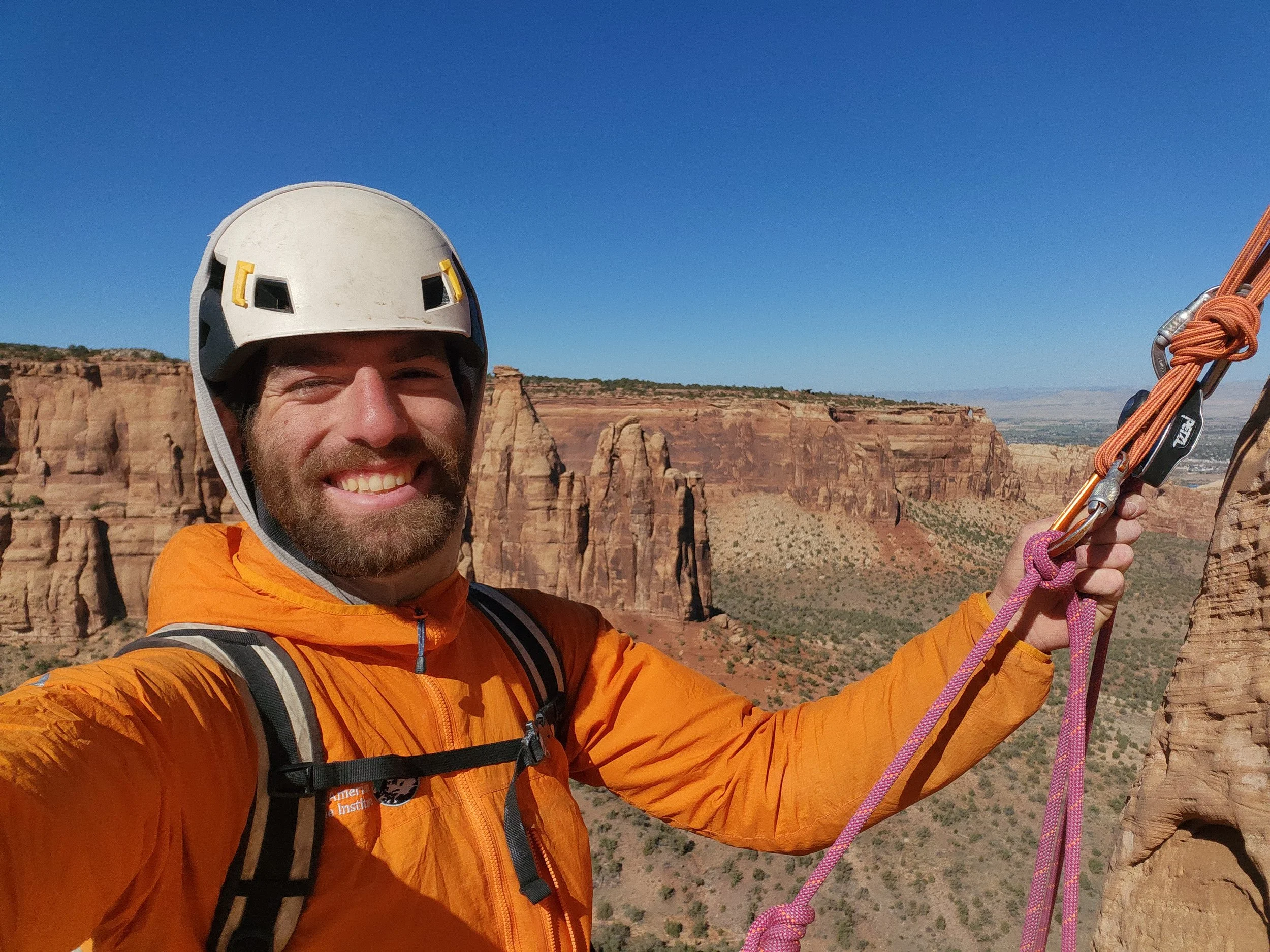 A smiling man in an orange jacket and a white helmet is taking a selfie with a scenic desert canyon landscape and rock formations in the background. He is holding onto climbing or rappelling equipment, including a pink rope and metal carabiners.