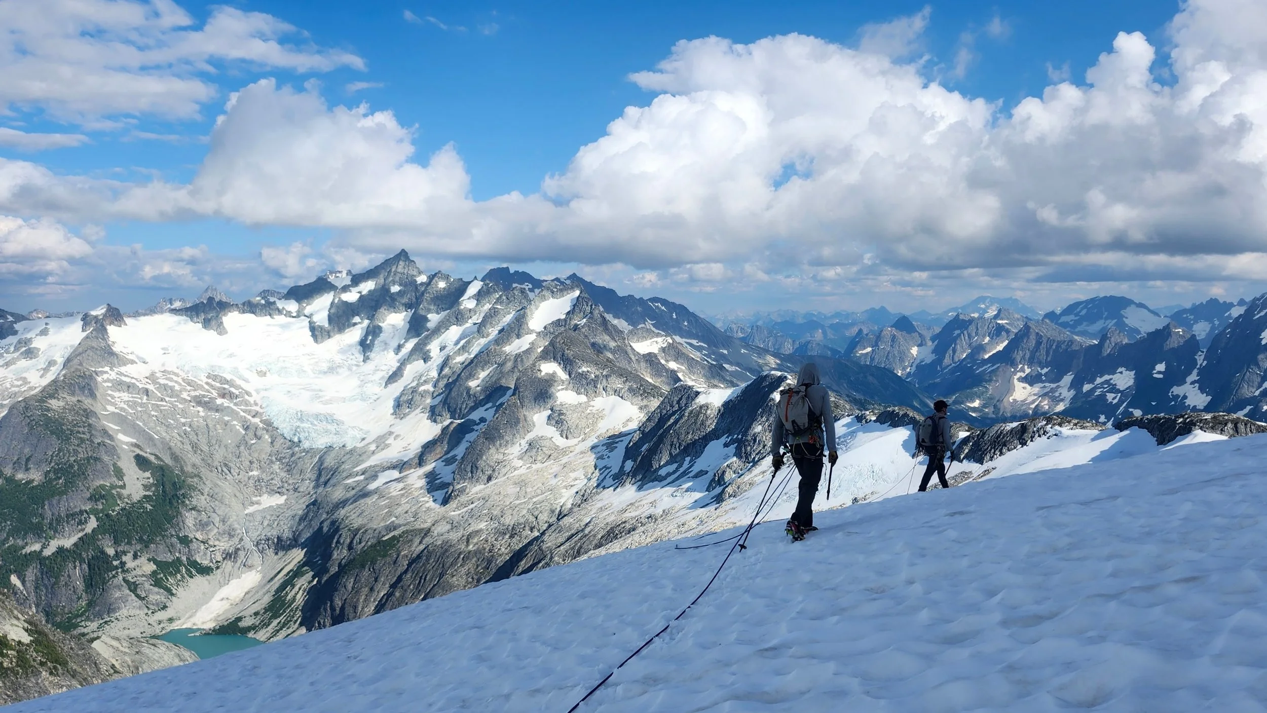 Two hikers with backpacks and trekking poles hiking on snow-covered slopes in a mountainous landscape with snow-capped peaks, glaciers, and a partly cloudy sky.