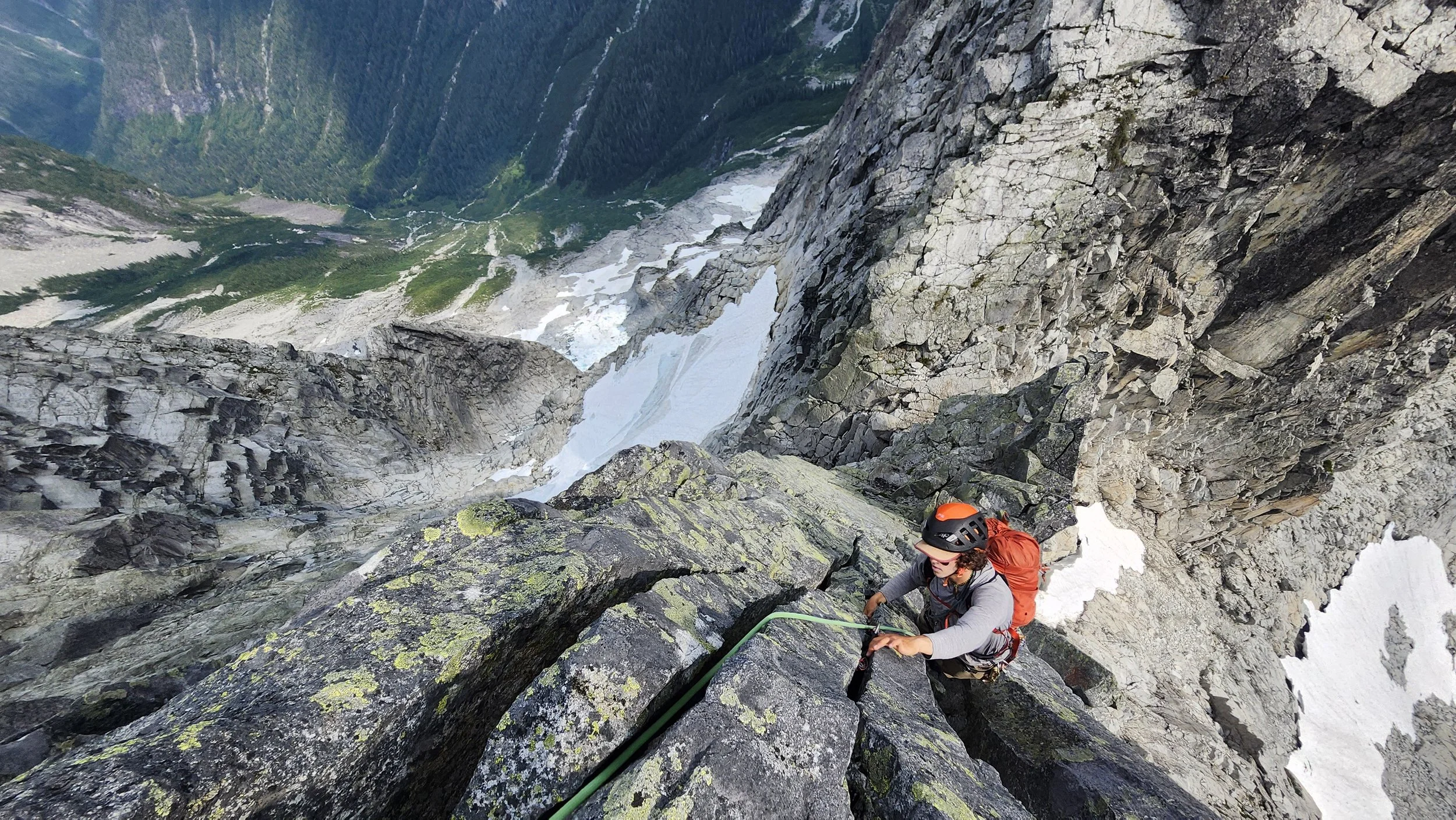 A person wearing a helmet and orange backpack climbing a steep mountain with rugged rocks and snow, overlooking a lush green valley with trees and mountains in the background.