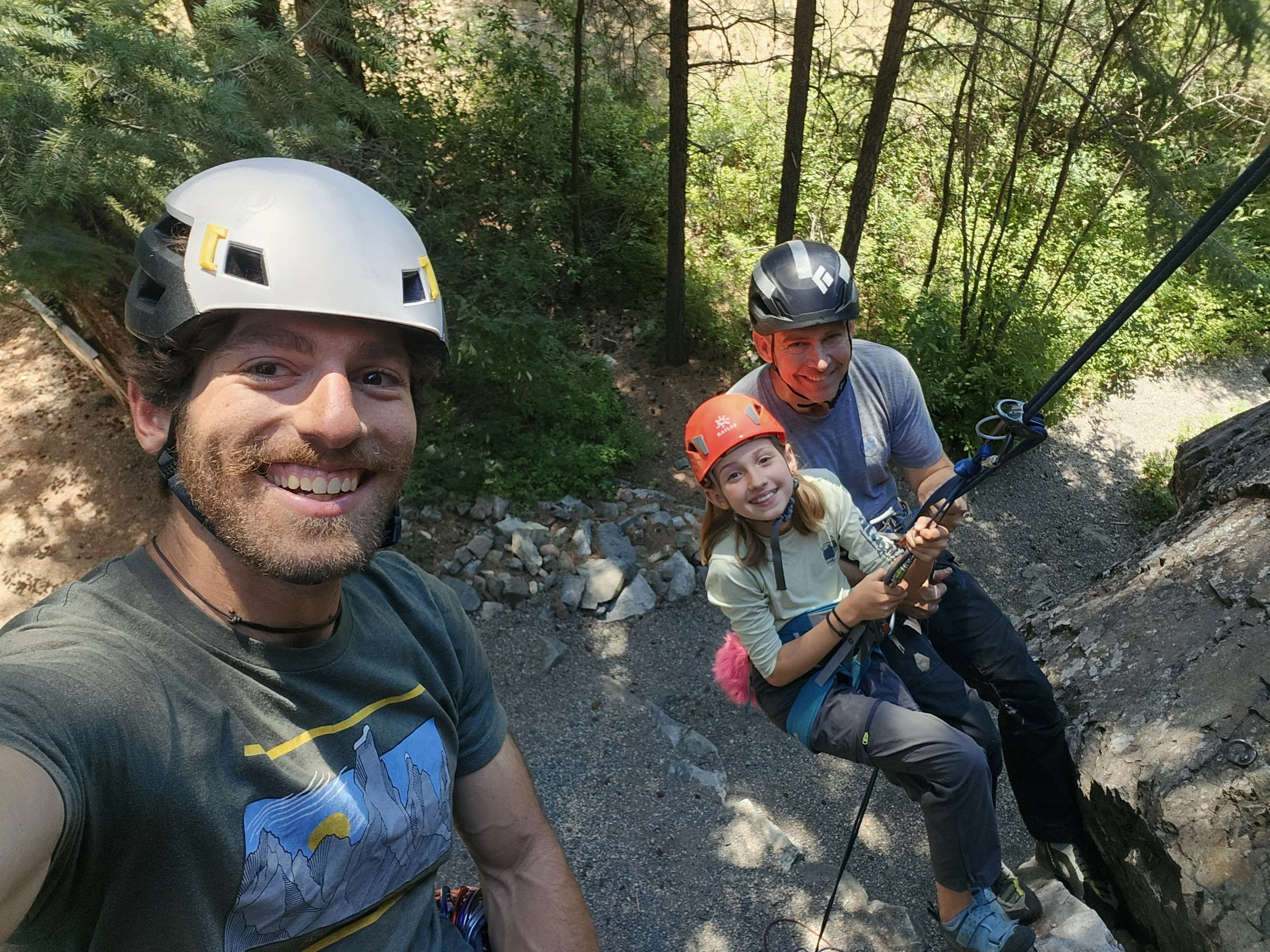 Four people wearing helmets, including two adults and two children, smiling and posing for a selfie outdoors in a wooded area while climbing a rock face.