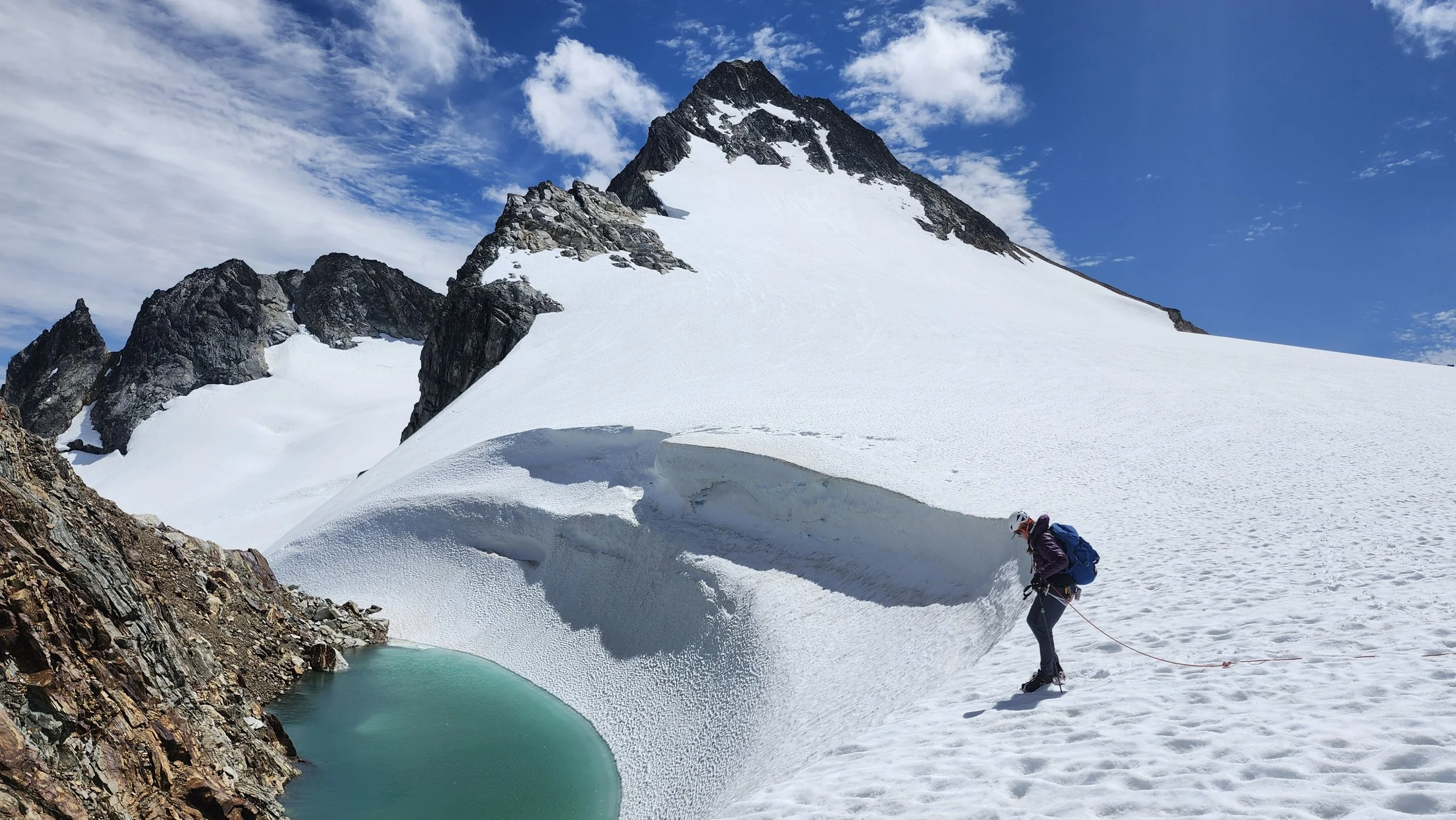 A person wearing a helmet and backpack hikes on snow-covered terrain near a turquoise glacial lake, with a steep, snow-covered mountain and rocky outcroppings in the background under a partly cloudy blue sky.