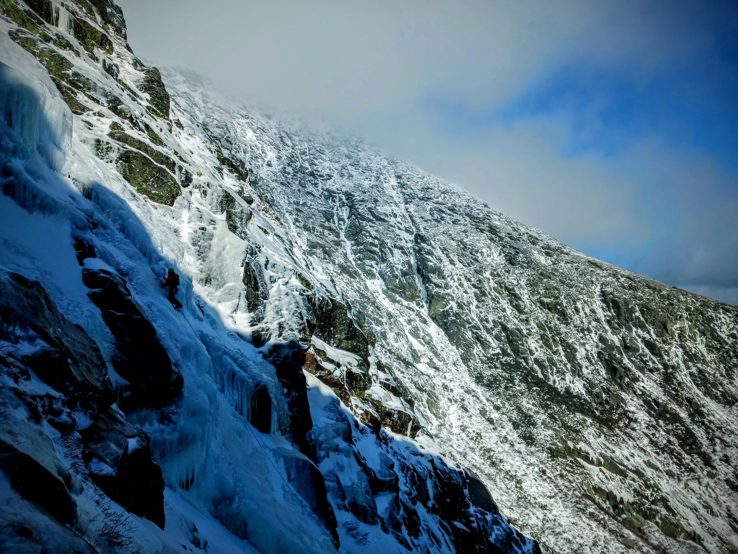 A person ice climbing on a steep, icy mountain slope covered in snow and ice, with rugged rocky terrain and a partly cloudy sky in the background.