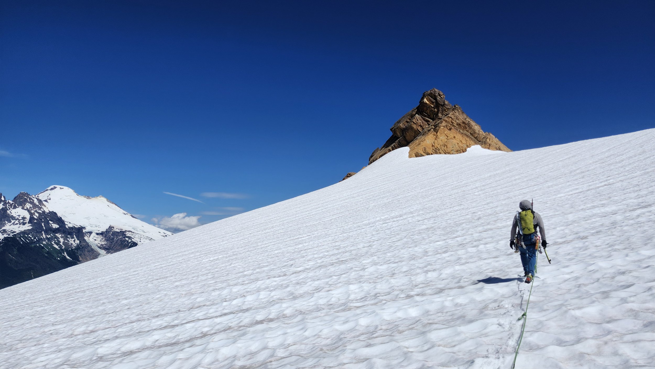 A person wearing outdoor gear and a yellow backpack climbing a snow-covered mountain slope with rocky peaks in the background under a blue sky.