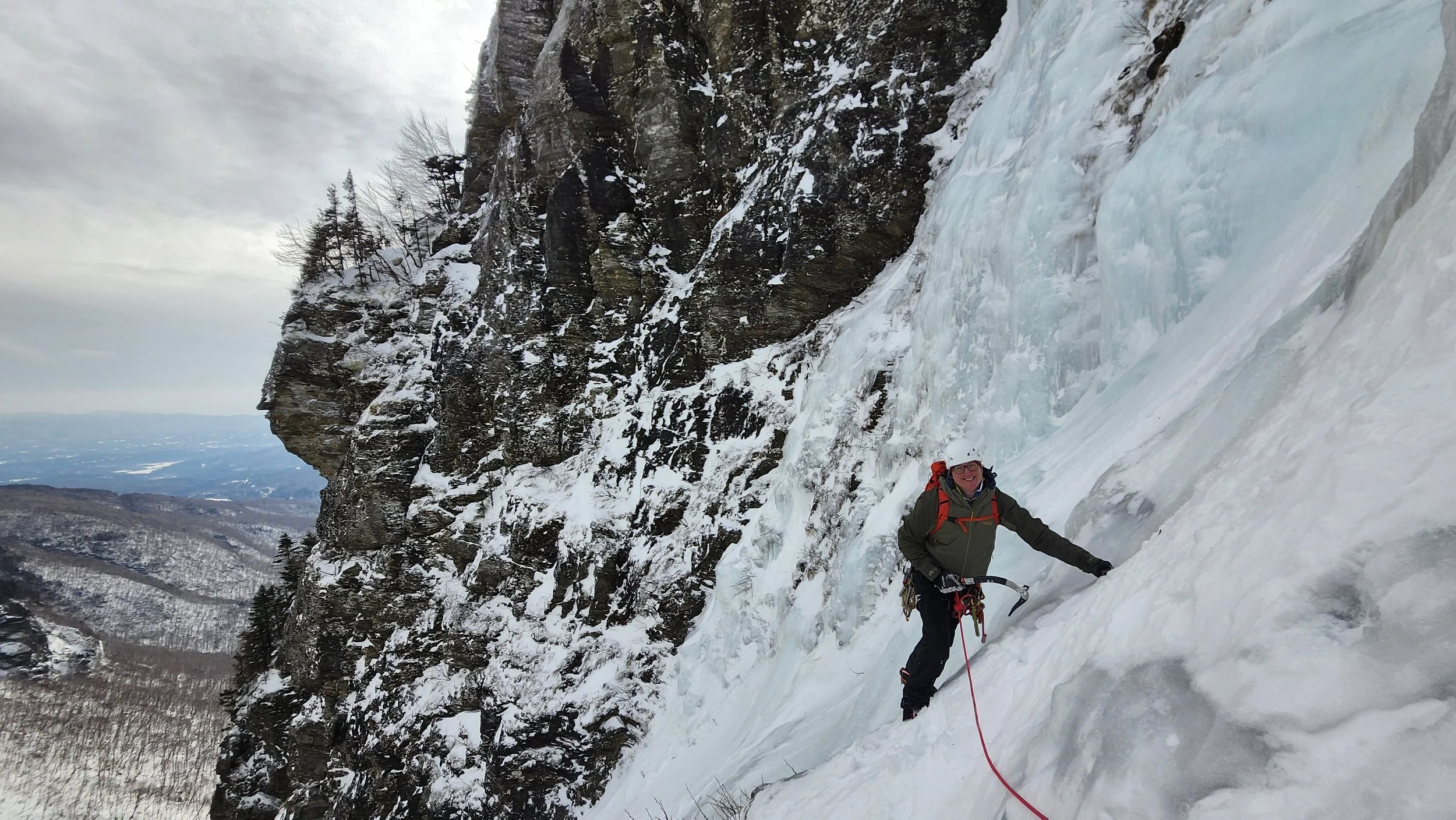 A person in climbing gear standing on a snowy and icy mountainside, with a steep cliff face and leafless trees in the background, holding a climbing tool and smiling.