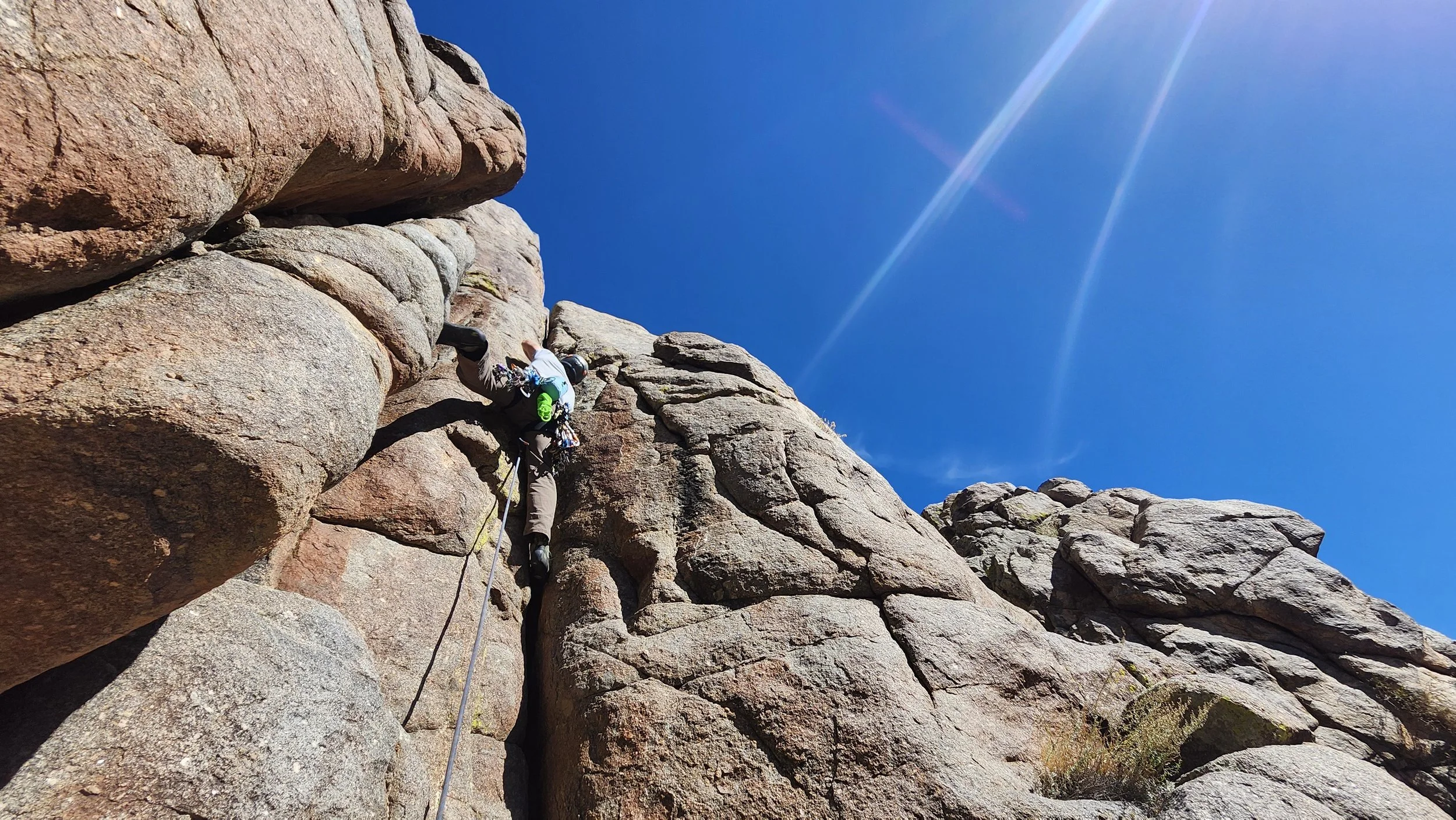 A person rock climbing on a steep rock face under a clear blue sky with a bright sunbeam.