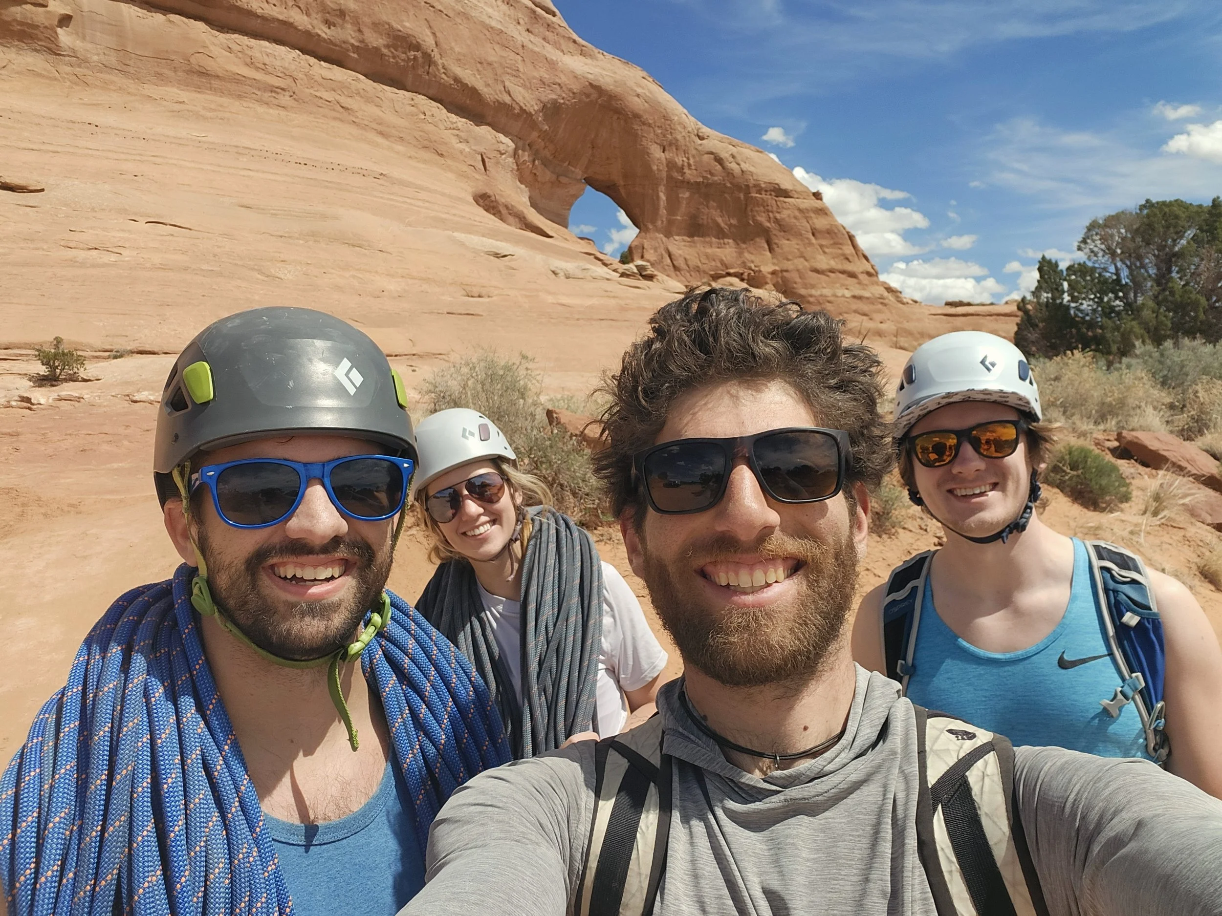 A group of five smiling people wearing helmets and sunglasses taking a selfie outdoors in front of a red rock canyon landscape with a rock arch formation under a blue sky with scattered clouds.