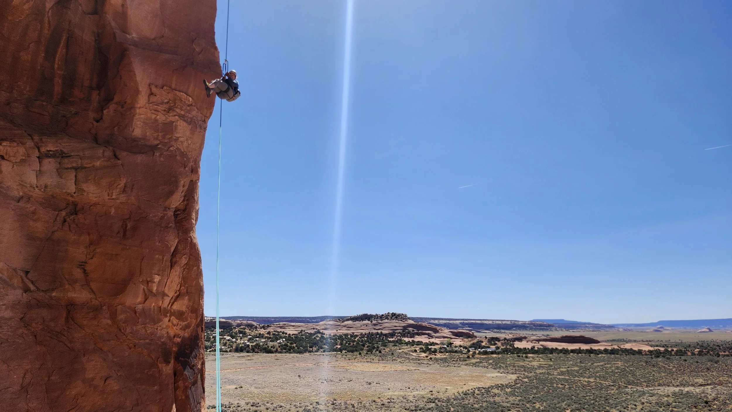 A person rappels down a red rock face in a desert landscape under a clear blue sky with visible jet trails.