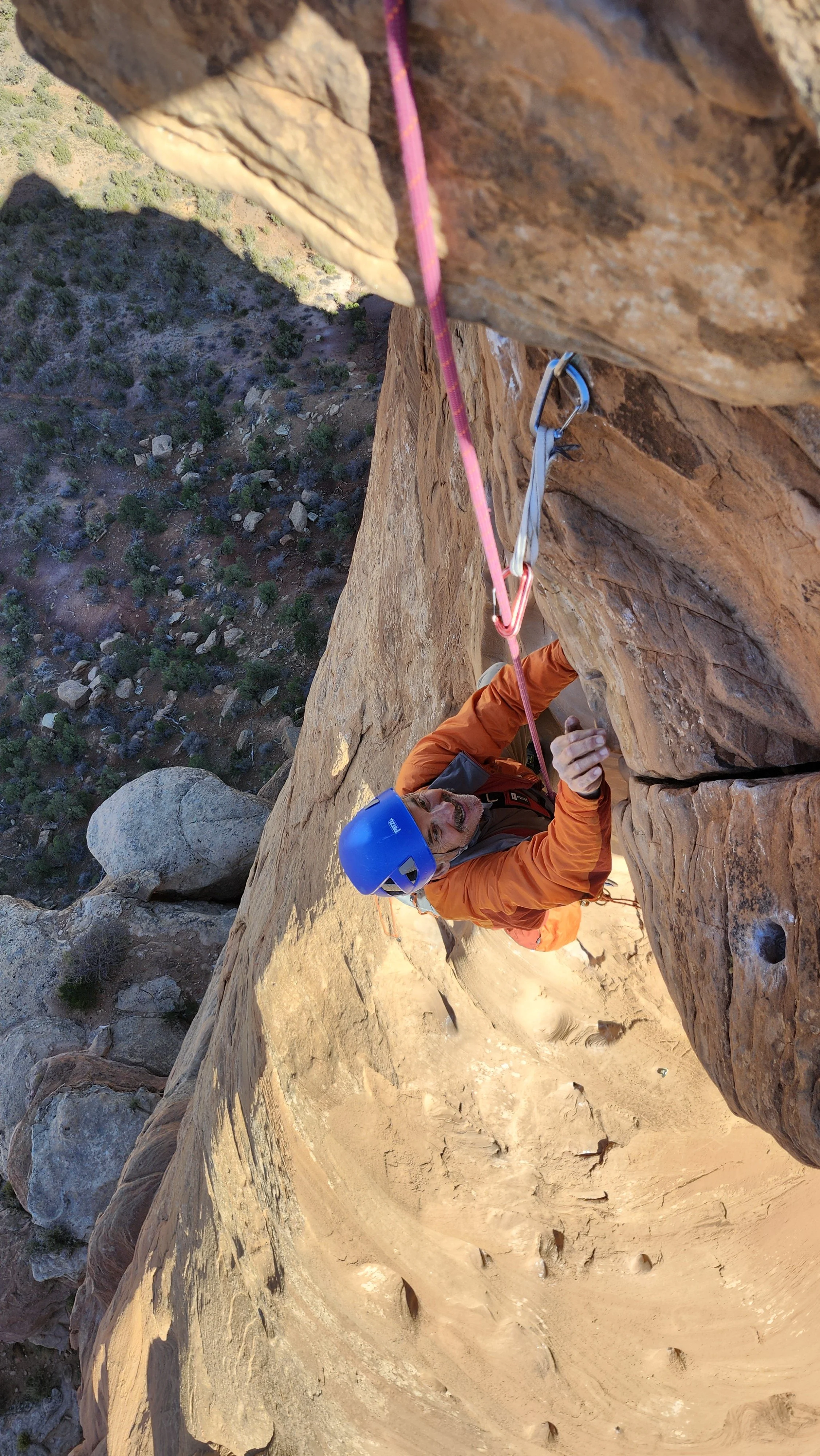 A person climbing a steep rock face with safety gear, including a blue helmet, orange jacket, and climbing harness, secured with a carabiner and pink rope. The landscape below shows a desert terrain with scattered rocks and vegetation.