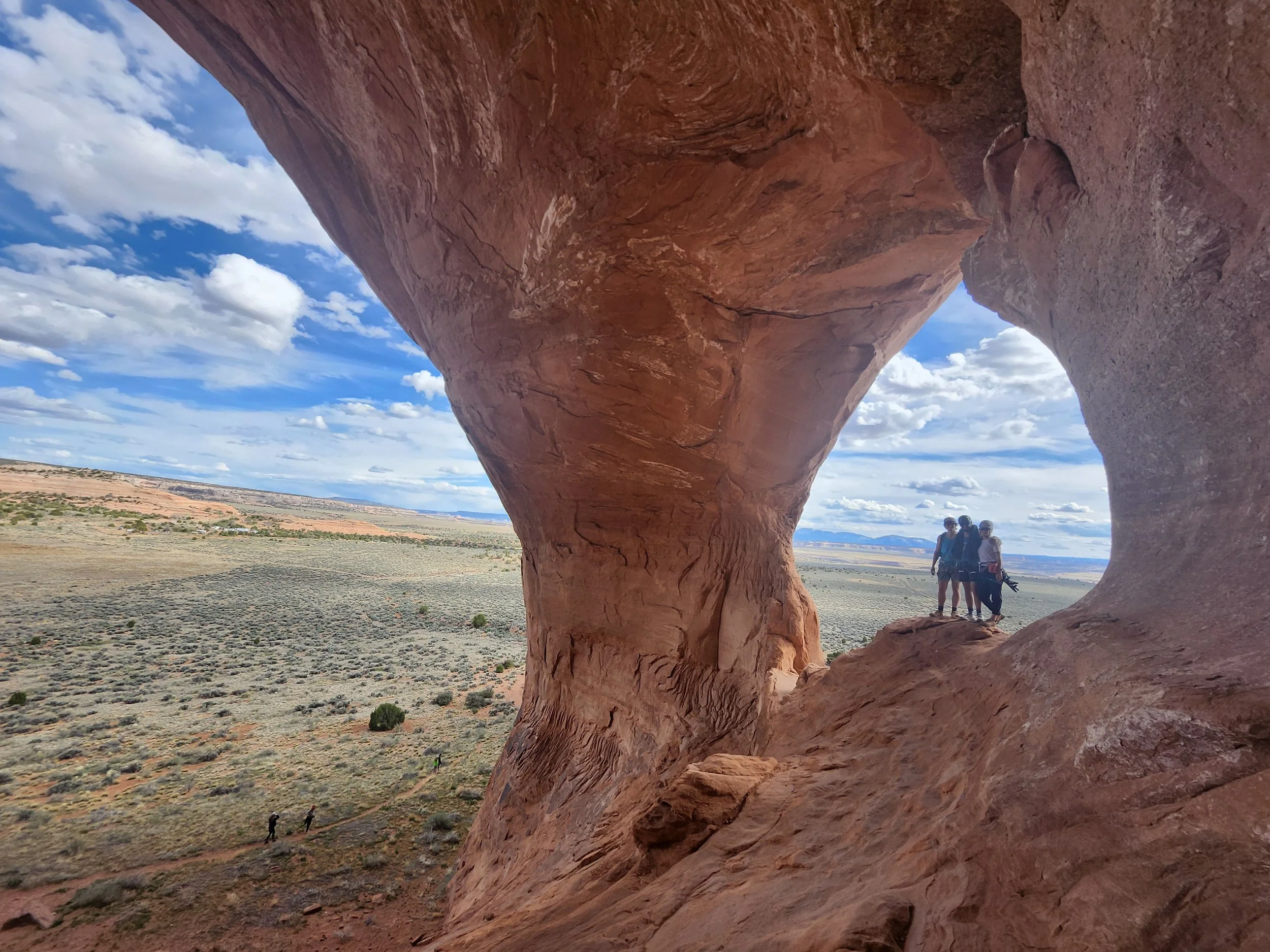 Red sandstone arches in a desert landscape with a blue sky with clouds, and a few people standing and walking in the distance.