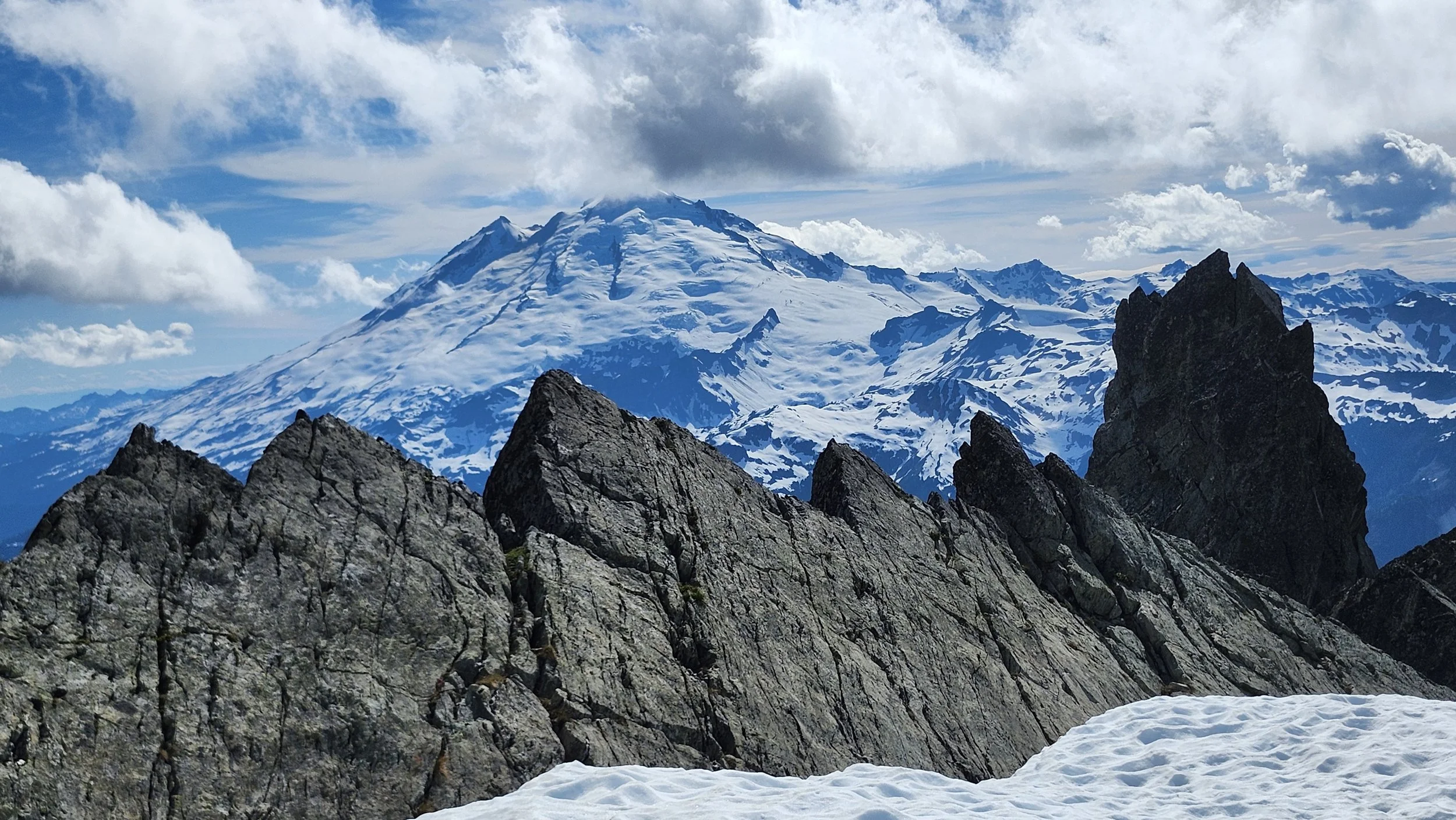 Snow-capped mountain behind rugged dark rock formations with a cloudy blue sky overhead.
