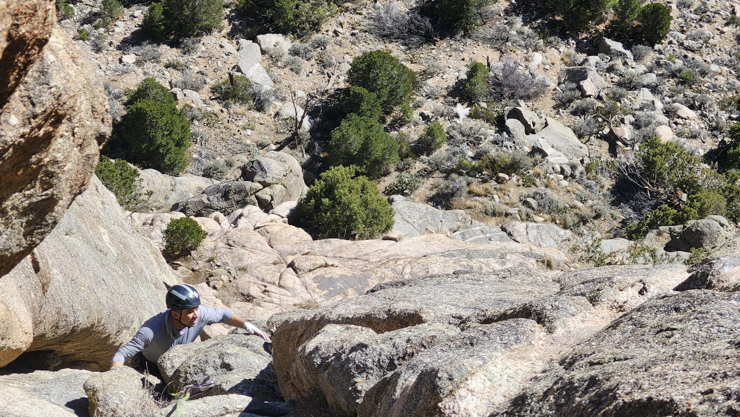 A person wearing a helmet climbing up a steep rock face in a mountainous area with rocky terrain and sparse vegetation.