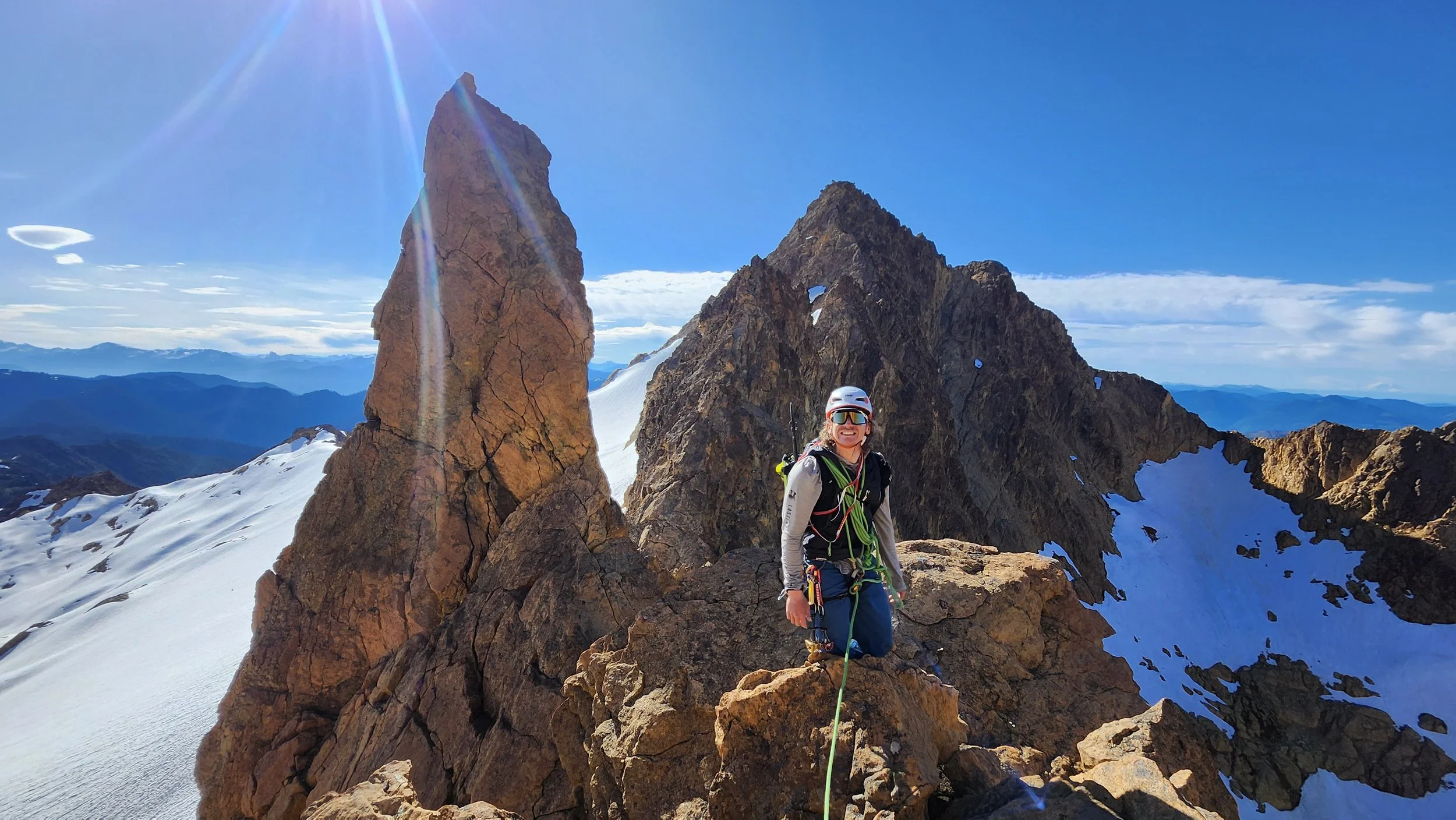 A person on a mountain summit wearing climbing gear, sunglasses, and a helmet, surrounded by jagged rocks and patches of snow, with mountain ranges and a clear blue sky in the background.