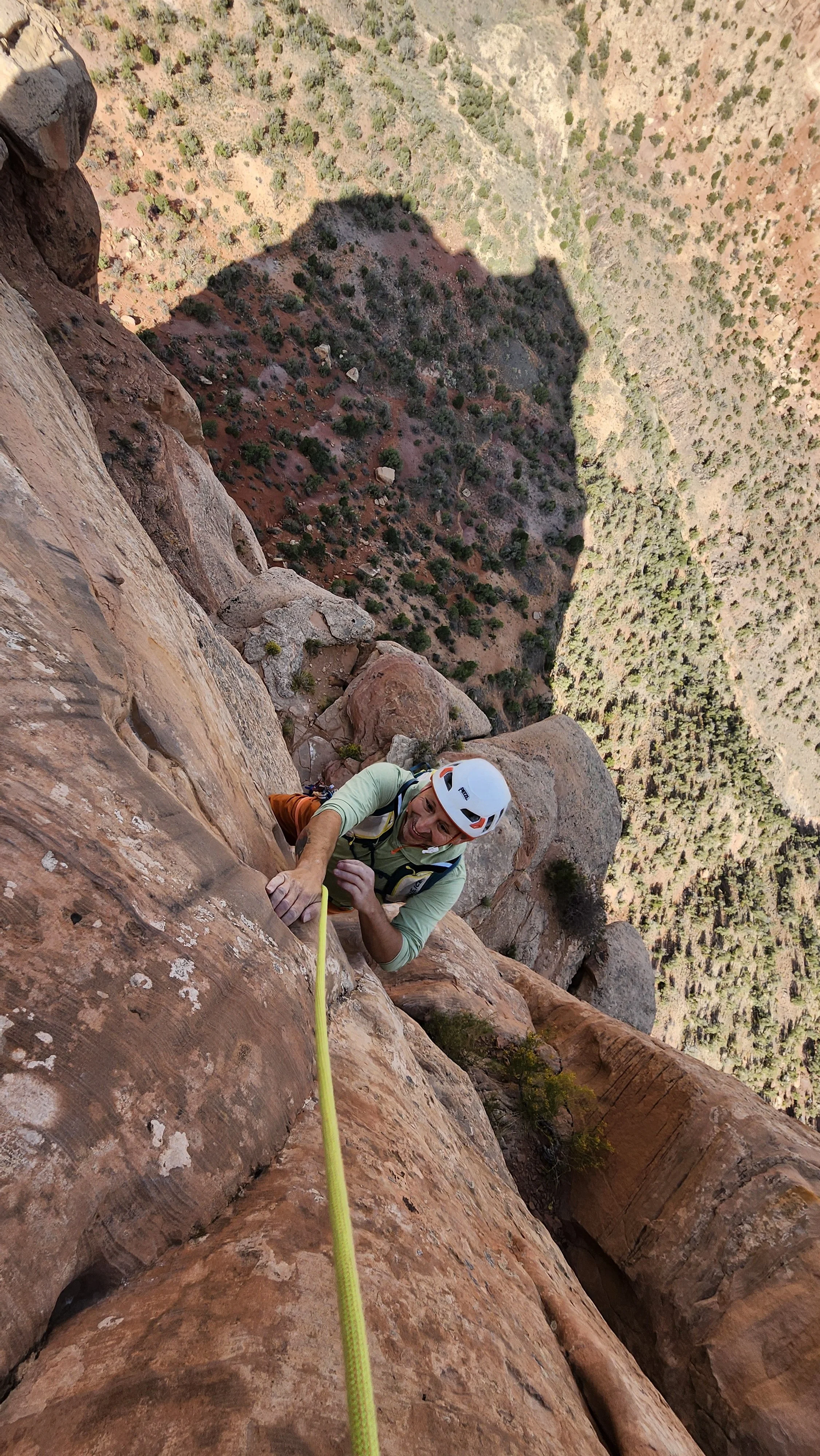 A man in climbing gear, including a helmet, is rock climbing on a steep cliff with a bright yellow safety rope. He is smiling and looking up at the camera while gripping the rock face with his hands. The background shows a deep canyon with sparse vegetation and rocky terrain.