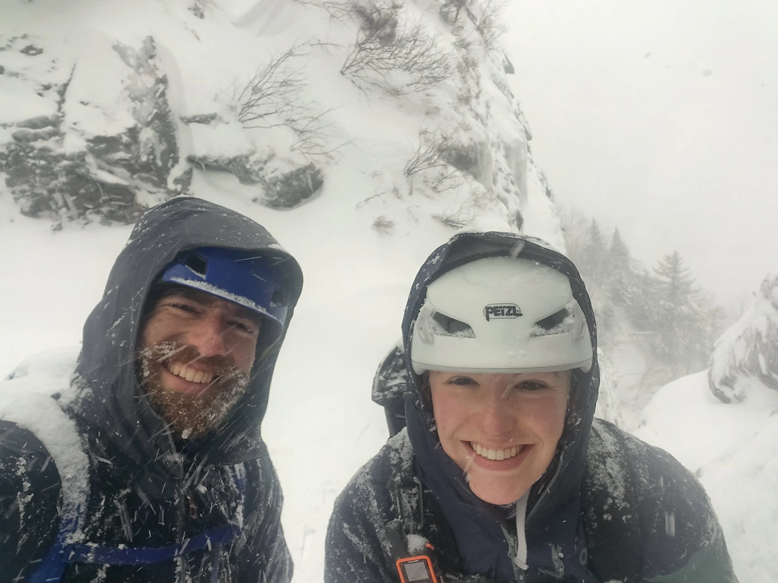 Two smiling people in winter gear taking a selfie outdoors in a snowy landscape with snow-covered trees and rocks.