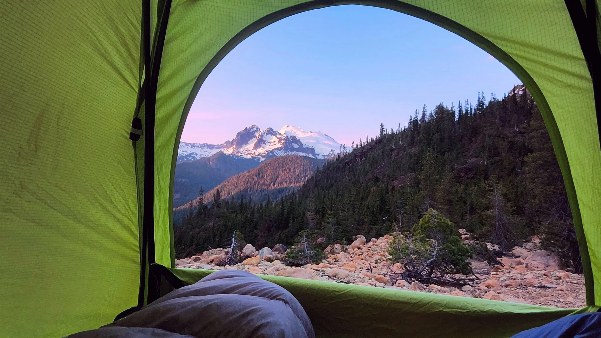 View from inside a bright green tent showing snow-capped mountains and evergreen trees with a rocky foreground.