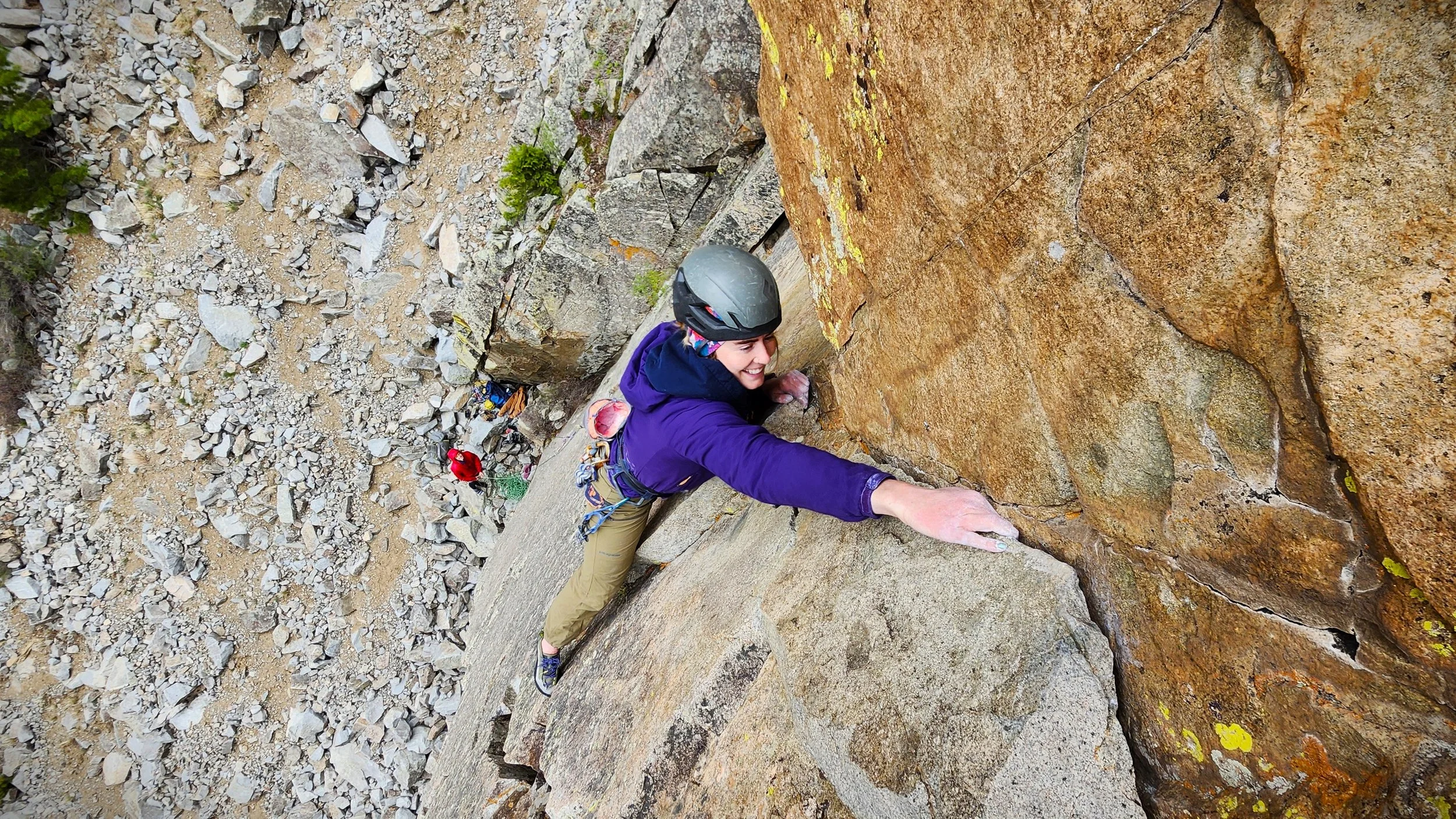 A person rock climbing on a steep, brownish-orange rock face, wearing a gray helmet, purple jacket, and beige pants, smiling as they reach for a hold.