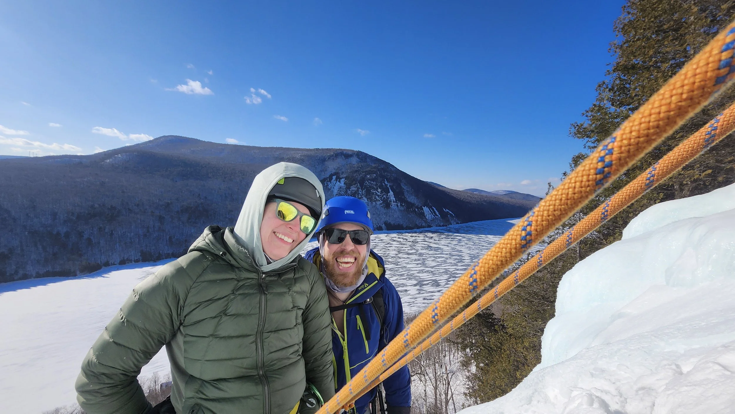 Two smiling people in winter gear and sunglasses taking a selfie on a snowy mountain, with mountains and trees in the background and a bright blue sky.