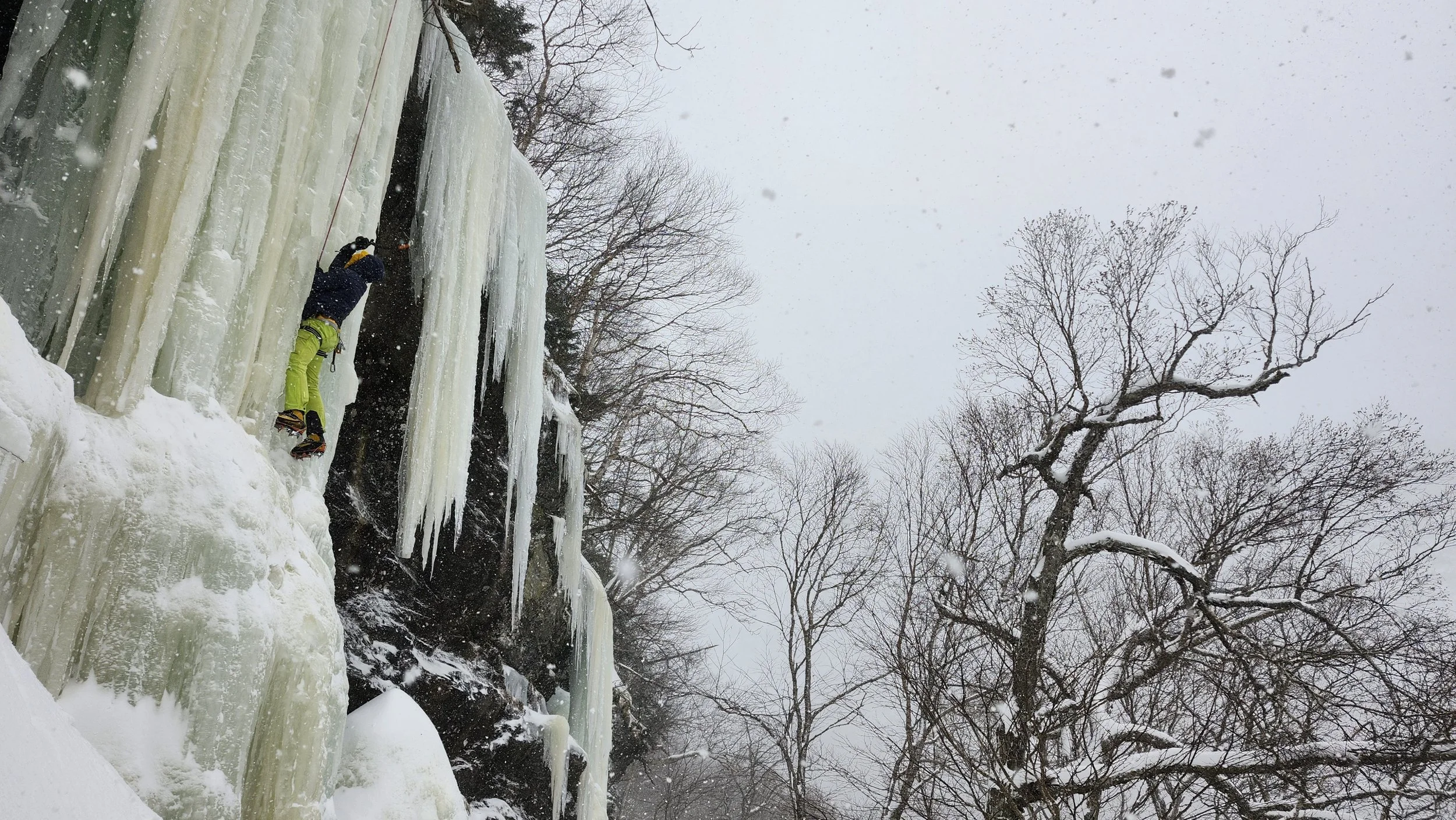 Person ice climbing on a frozen waterfall with snow-covered ground and leafless trees in the background.