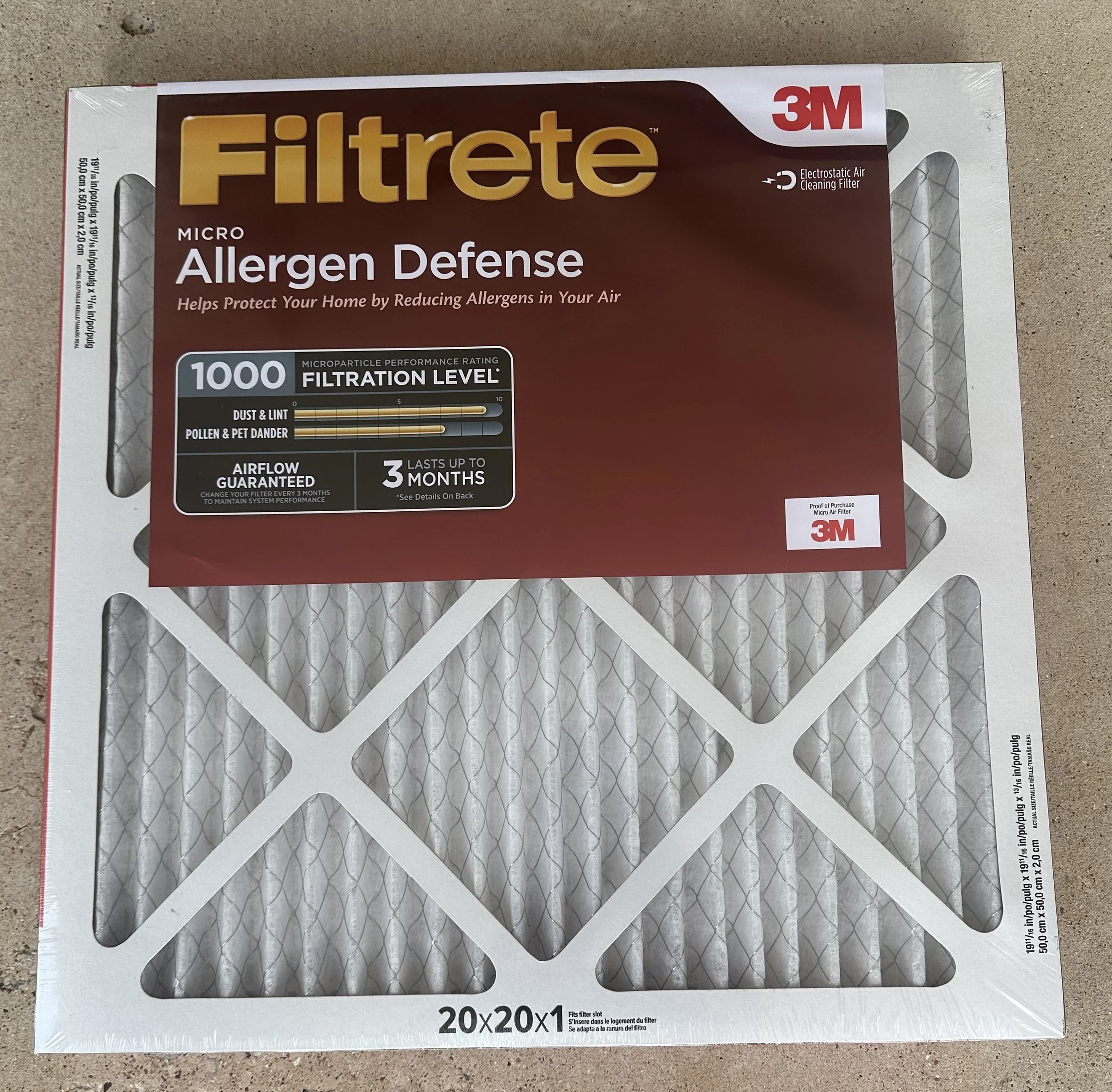 A box of 3M Filtrete Micro Allergen Defense air filter with dimensions 20x20x1 inches, resting on a gray surface. The box features labels and a brief description of its purpose to reduce allergens and improve air quality.
