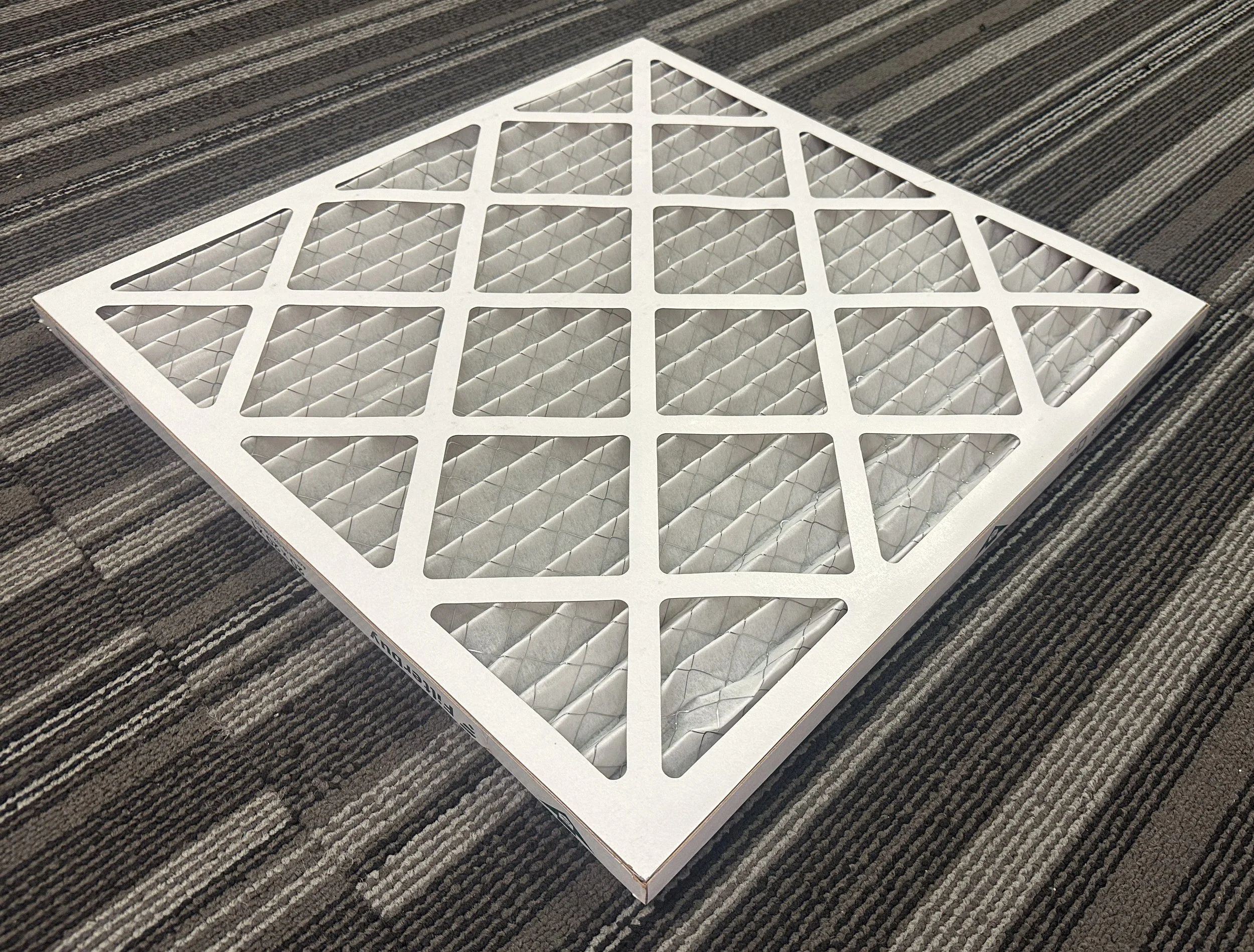 A rectangular white air filter with a diamond-shaped pattern, placed on a carpeted floor.