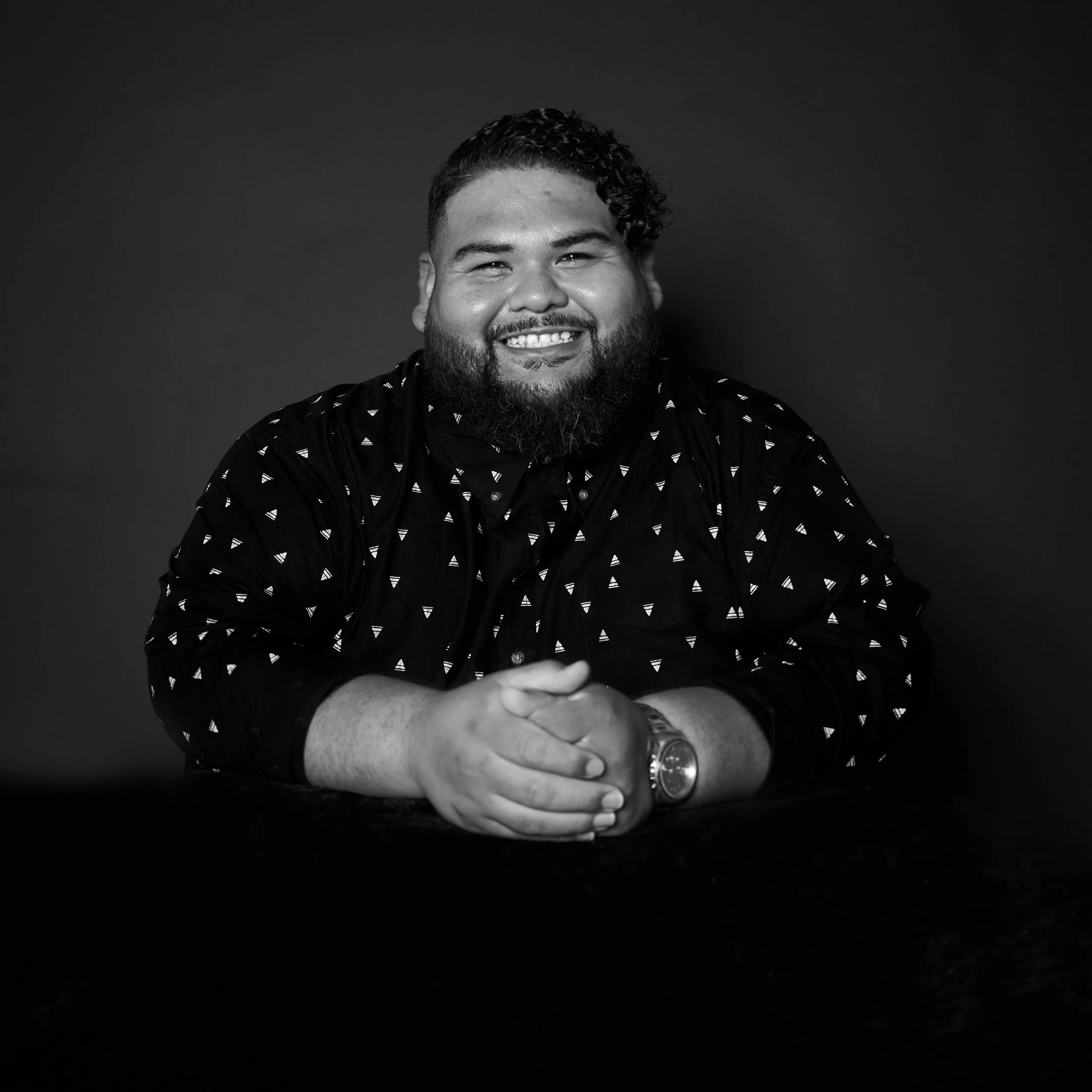 Portrait of a smiling man with a beard and short, curly hair, wearing a patterned shirt, against a dark background.